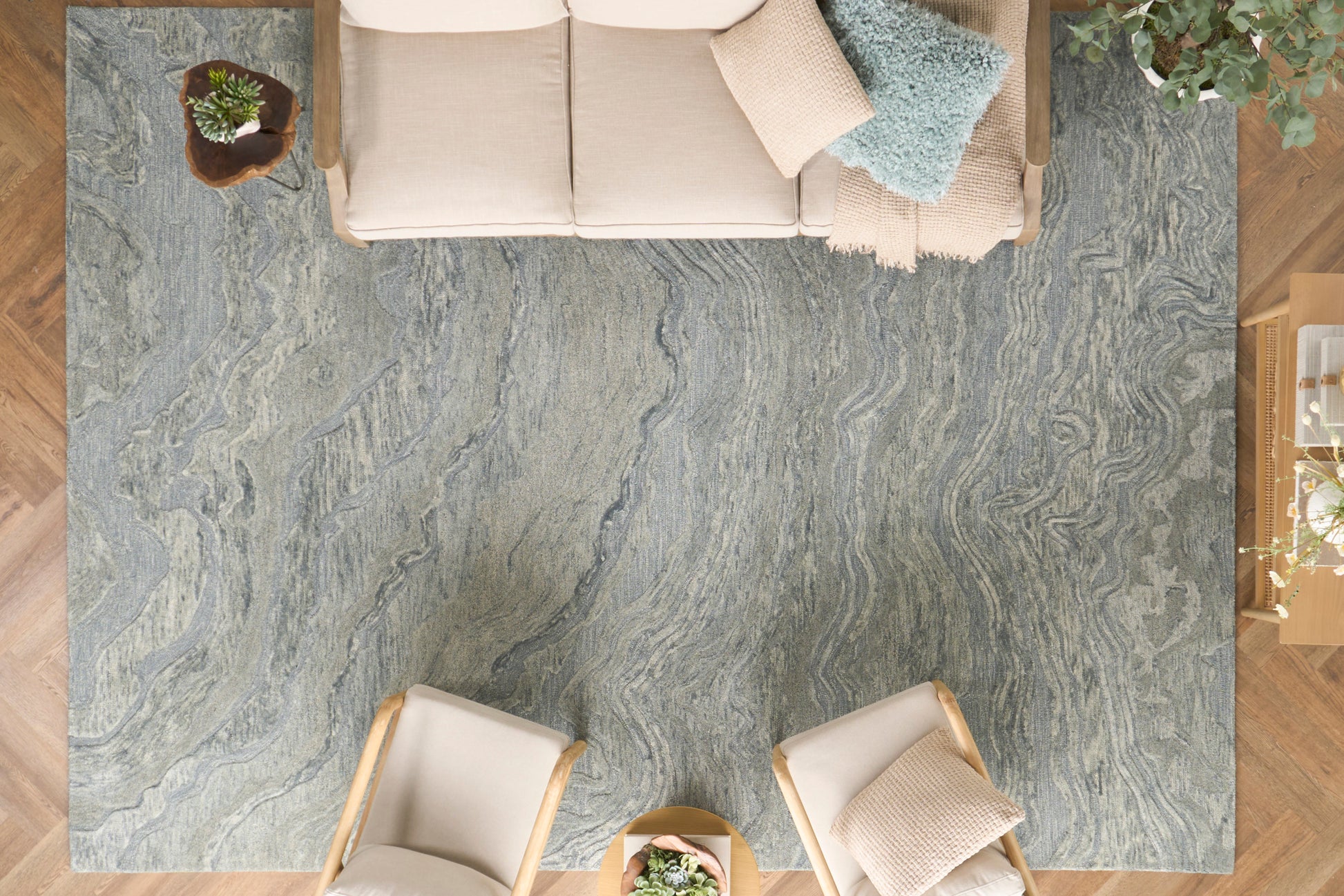 Overhead view of a blue-gray marbled area rug in a modern living room with a beige sofa & two wood-framed chairs, small round coffee table & plants.