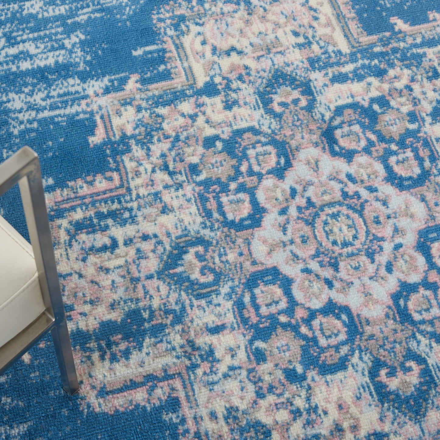 Blue Persian-pattern rug with pink floral medallions; metal-framed chair leg & white cushion in corner.