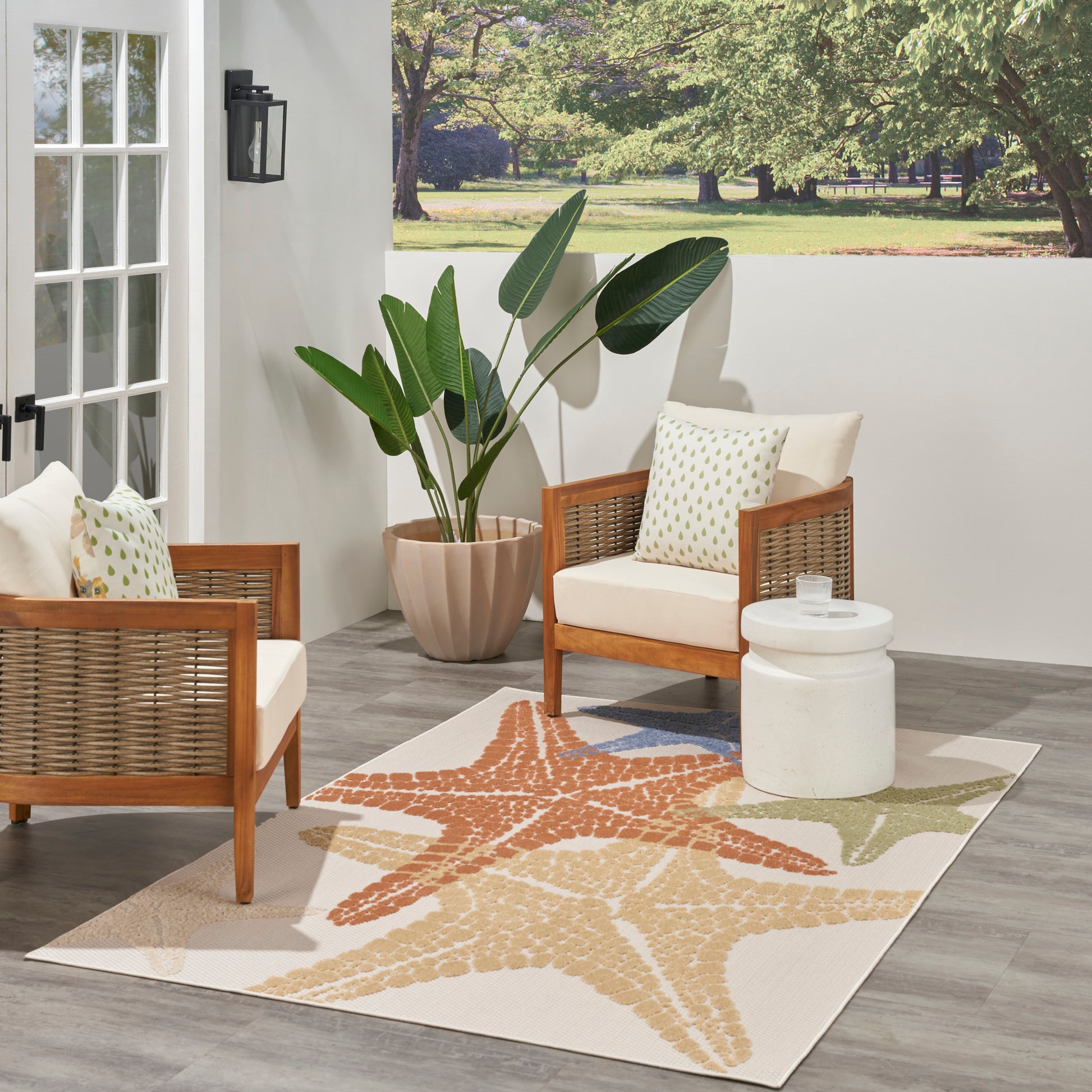 Outdoor rug with a starfish print (5' x 8') in a patio scene with cream cushions on wicker chairs, a white round side table, and a tropical plant in a terracotta pot.