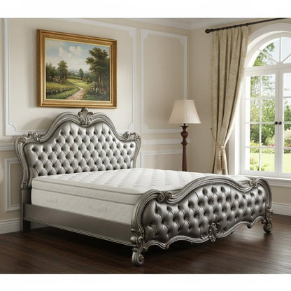 Luxurious silver tufted bed with ornate carved frame in a beige-paneled room, landscape painting, lamp, and large window.