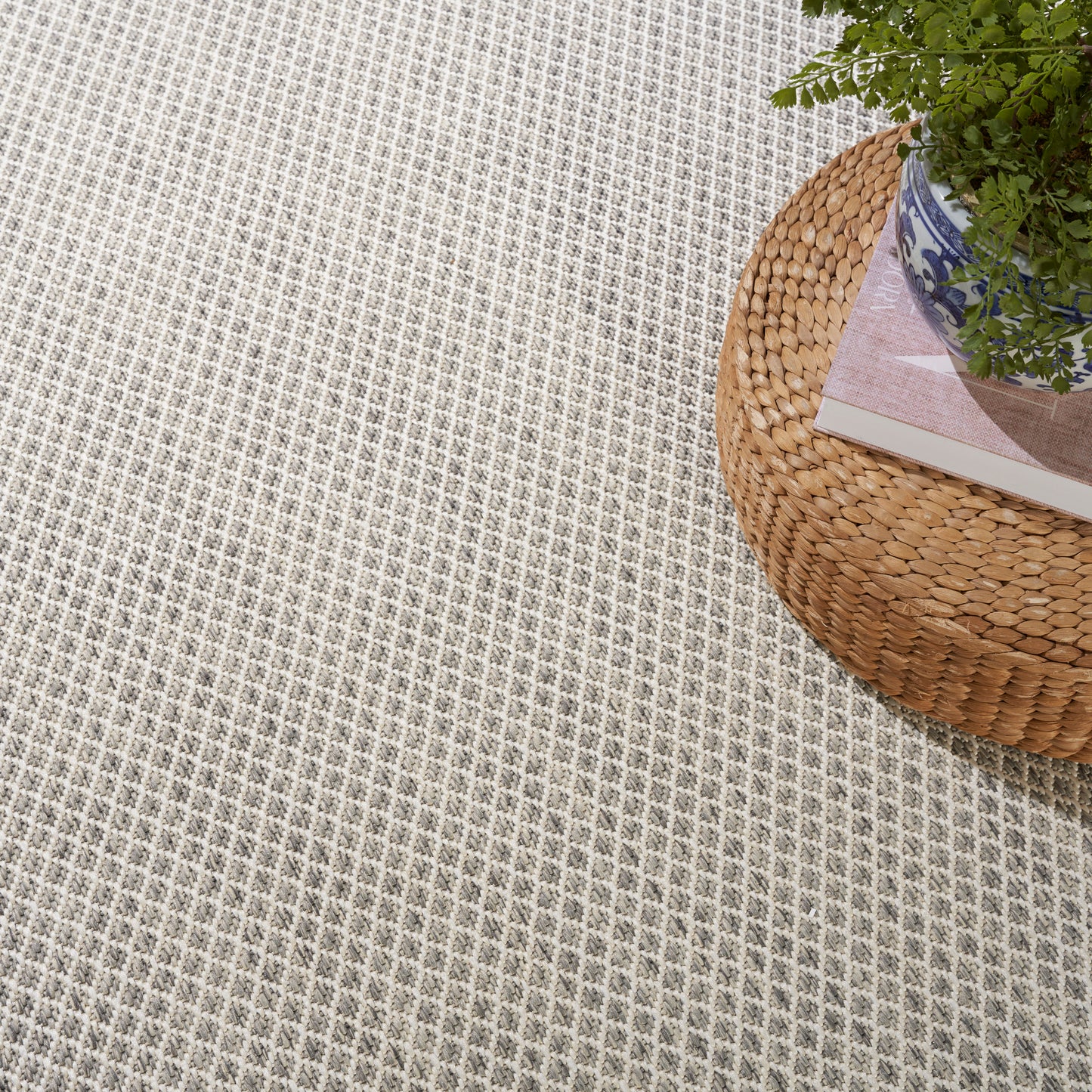 Beige textured woven rug with diamond grid pattern; round natural rattan ottoman topped with a plant and magazine.