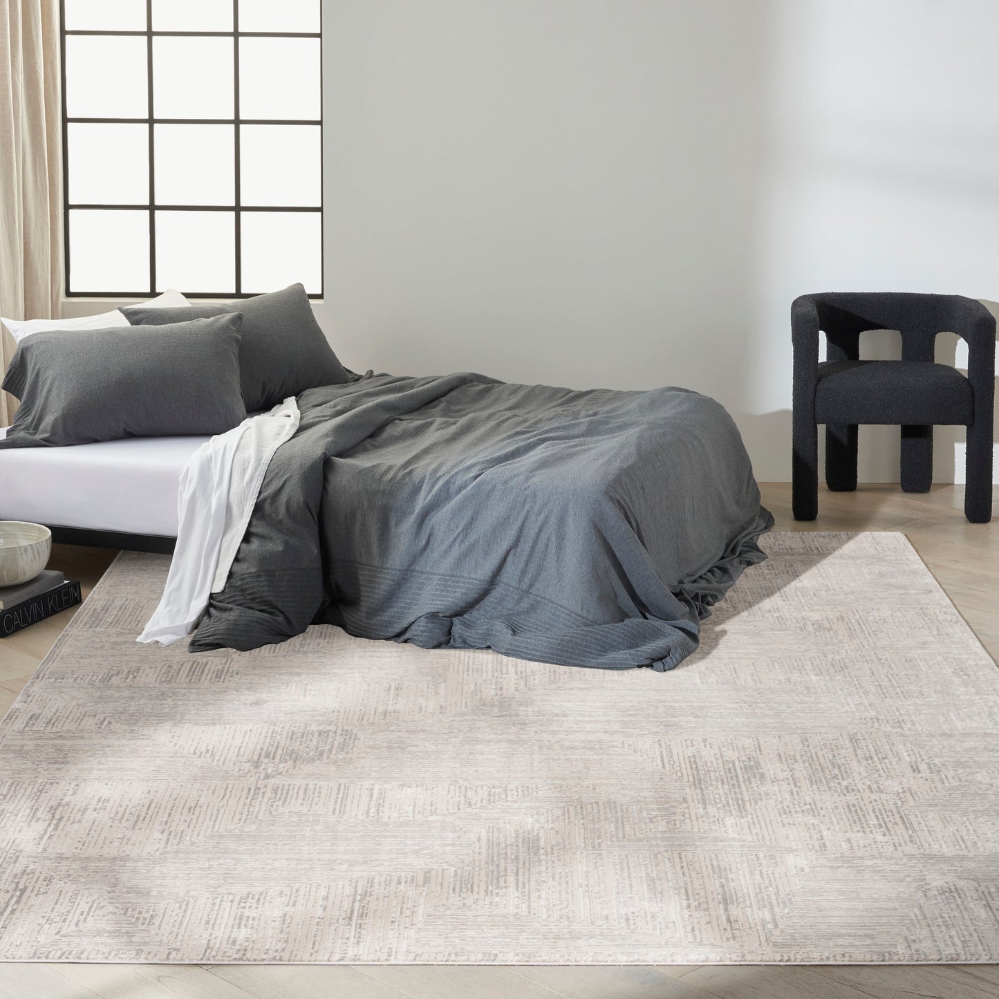 Beige textured rug on a minimalist bedroom floor with a dark gray duvet, white sheets, gray pillows on a low bed, and a black upholstered chair.