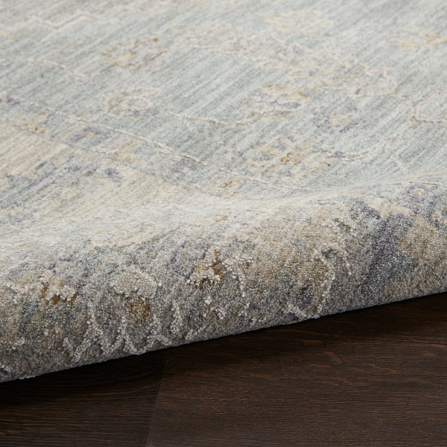 Soft blue-gray low-pile area rug with beige accents and a subtle damask pattern, rolled edge on dark wood floor.