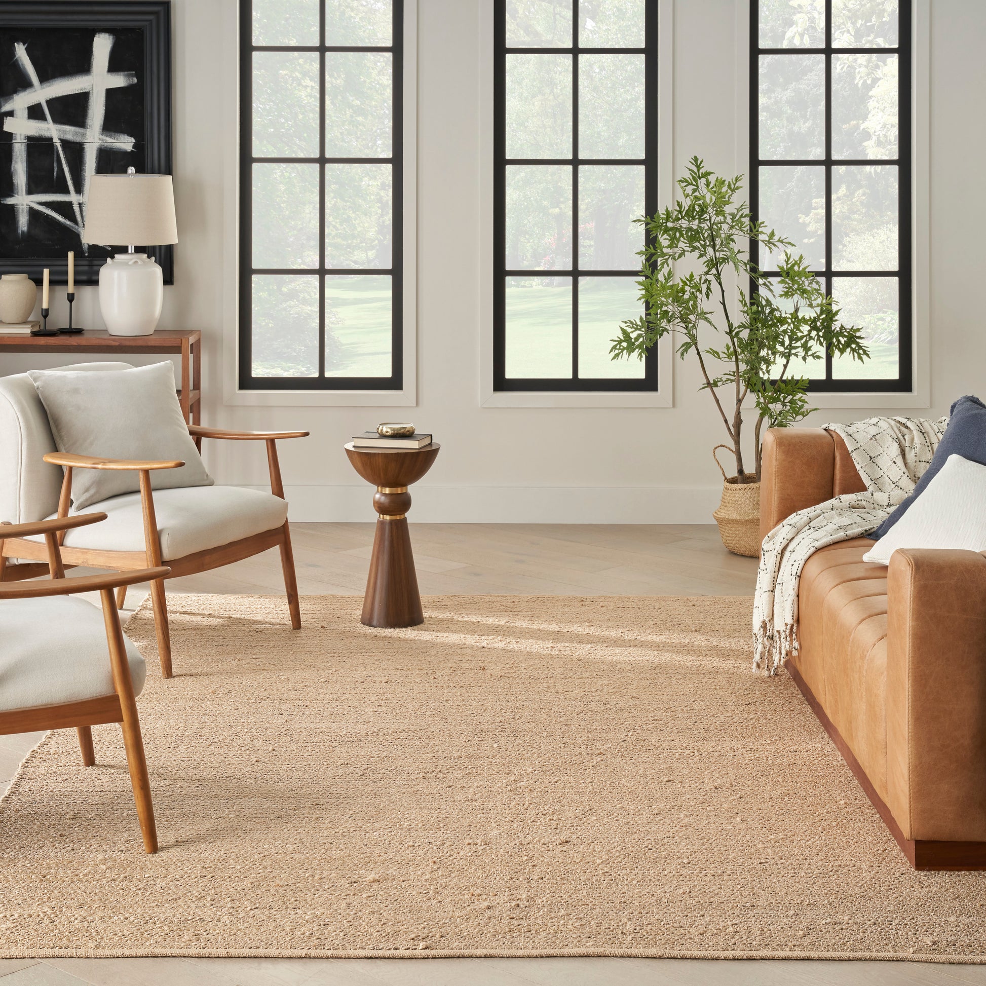 Bright living room with mid-century wood chairs, tan leather sofa, woven rug, large black-framed windows, potted plant.