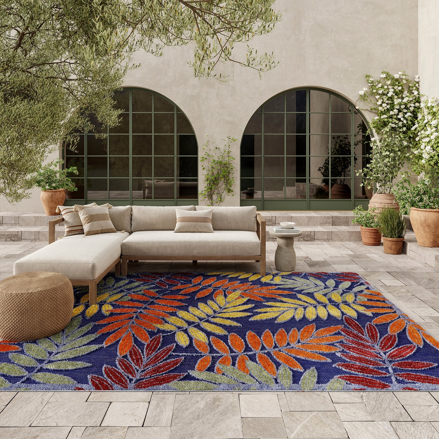 Outdoor patio with a blue leaf-pattern rug; beige sectional sofa, potted plants, arched windows in a light stucco wall.