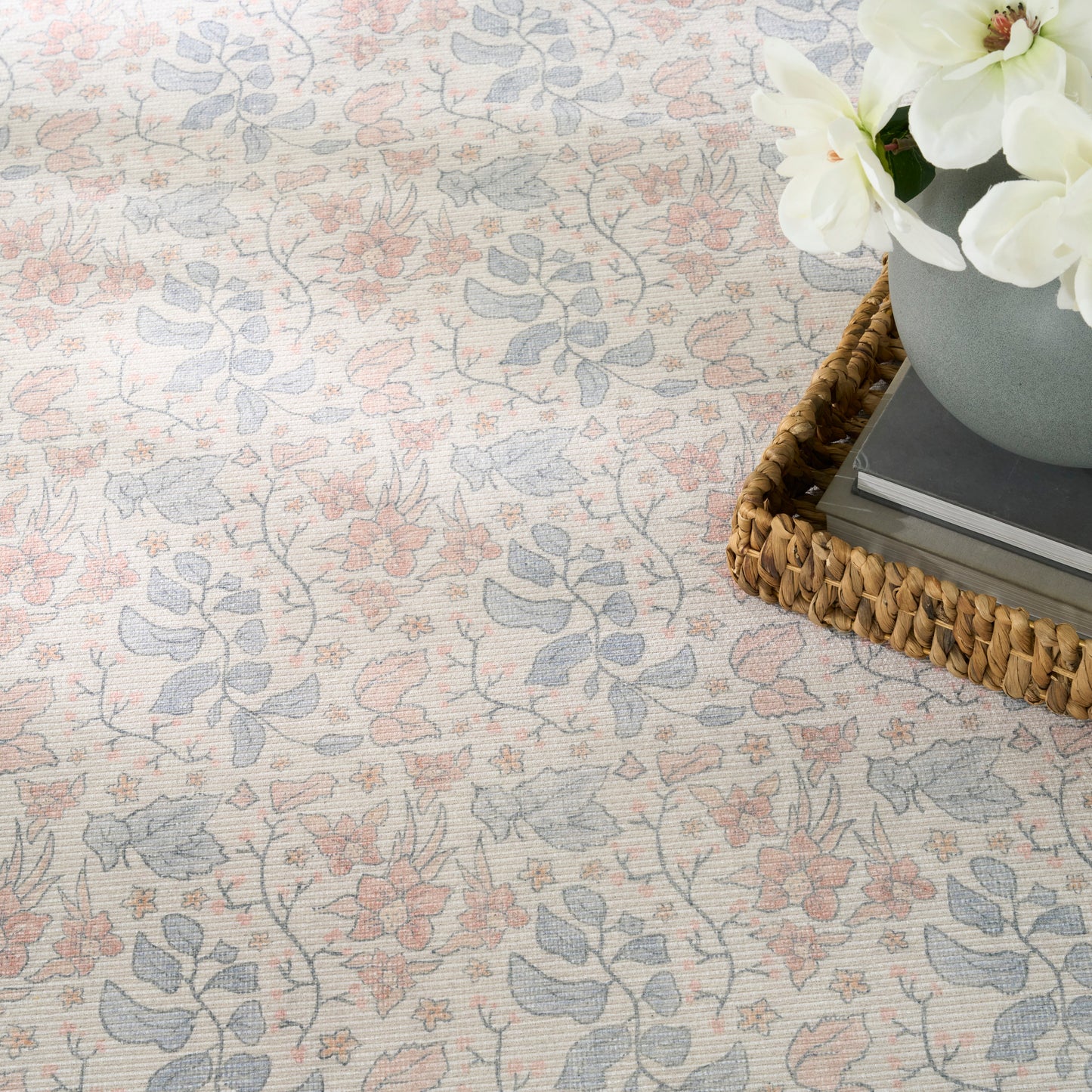 Pastel floral rug in pink and blue tones on beige; wicker tray with books and white flowers nearby.