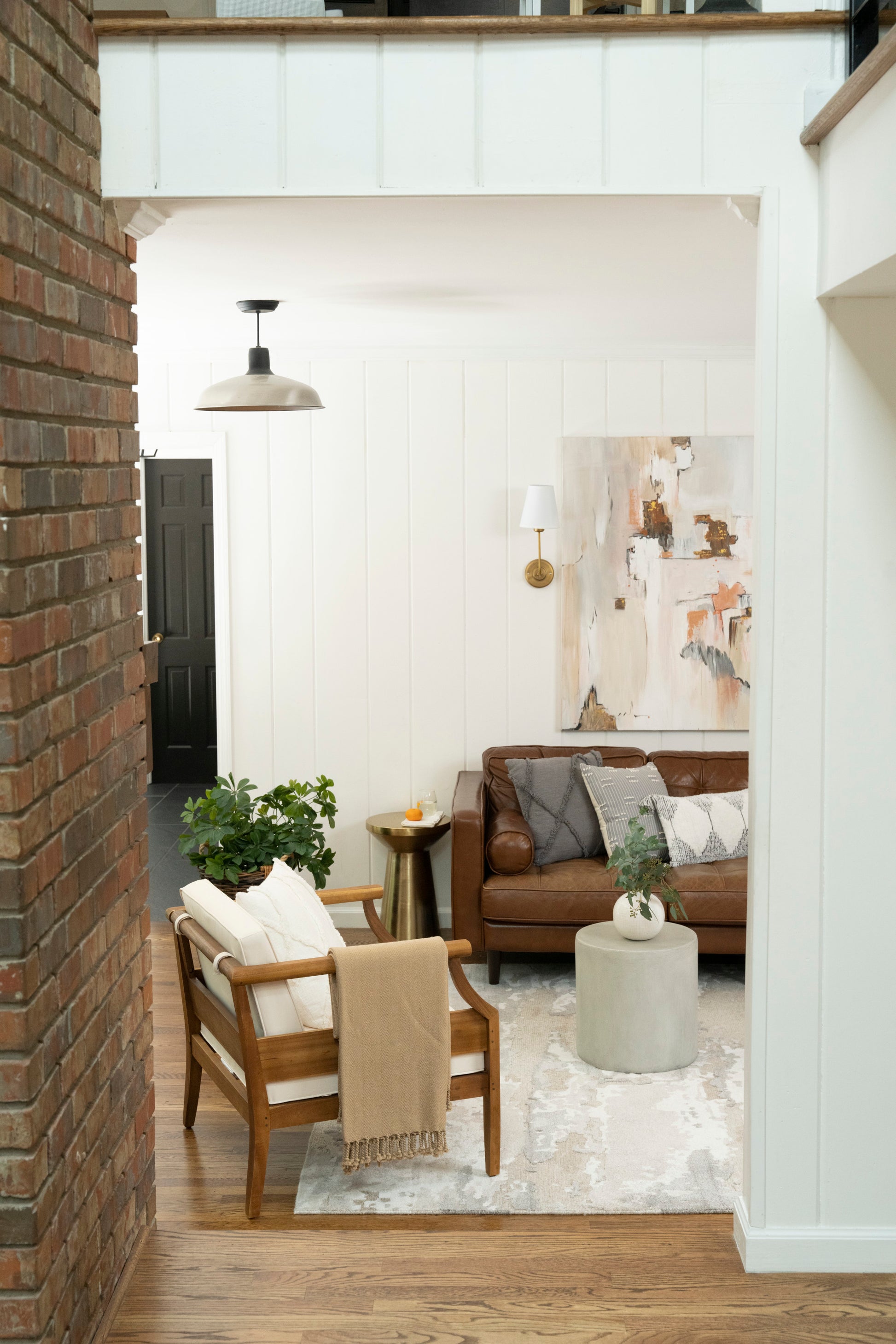 Warm living room with brown leather sofa, wooden armchair, beige throw, abstract art, white paneled walls and brick accent.