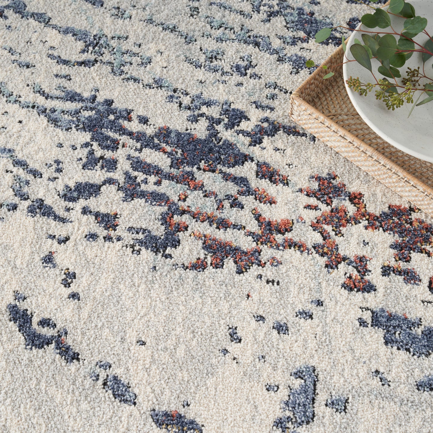 Cream rug with blue and rust abstract pattern; woven texture, wicker tray with greenery in the top-right.