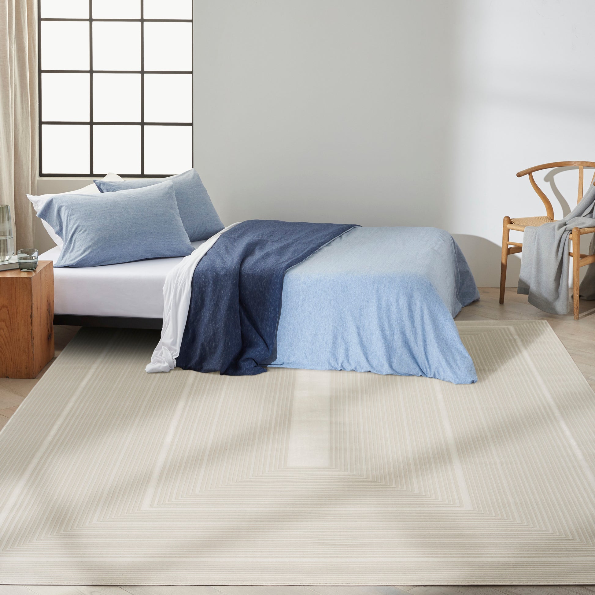 Light blue duvet on bed with navy throw, beige square-bordered rug, wooden nightstand & chair in sunlit modern bedroom.