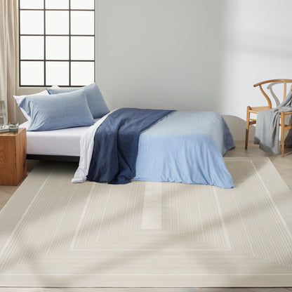 Light blue duvet on bed with navy throw, beige square-bordered rug, wooden nightstand & chair in sunlit modern bedroom.