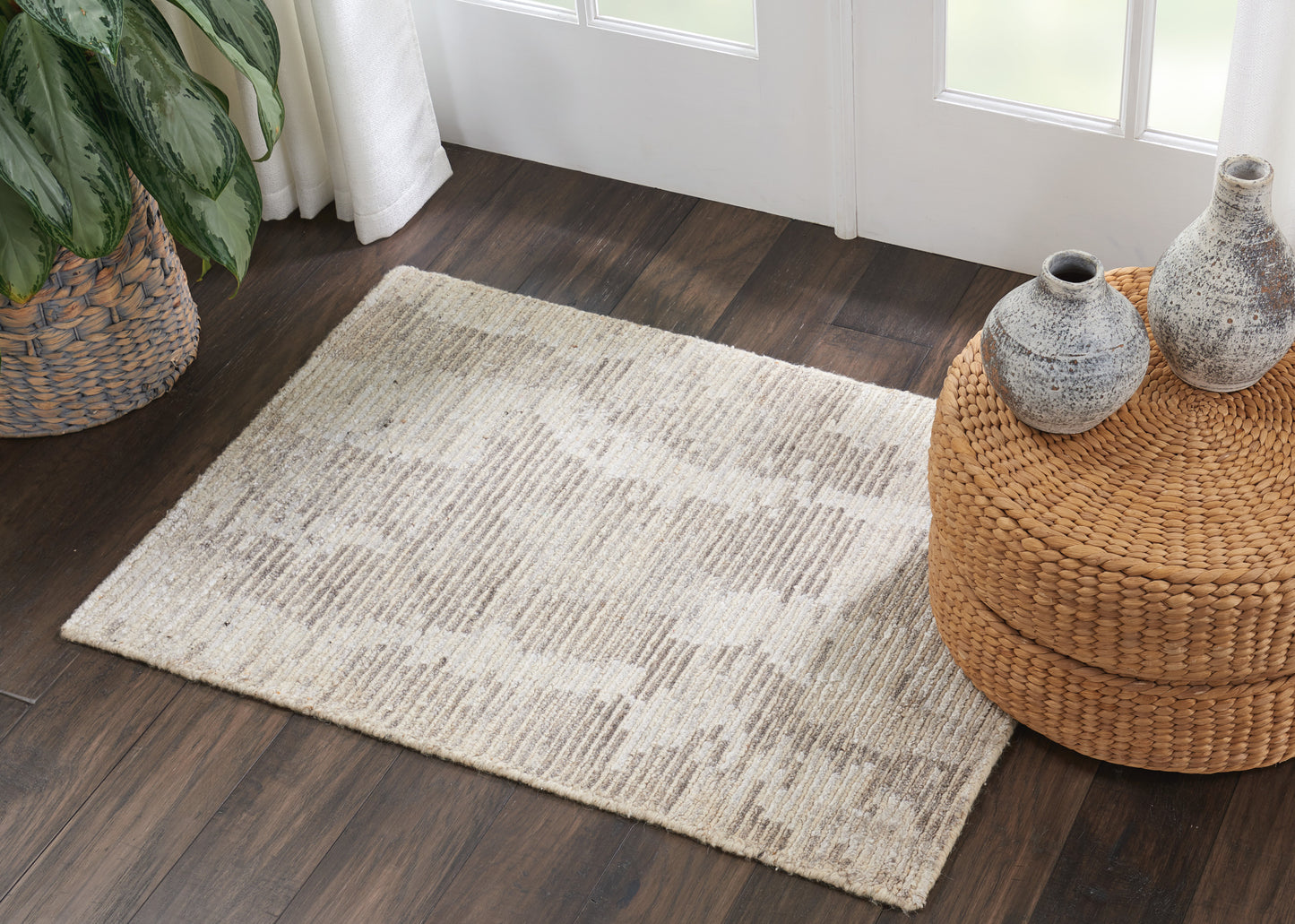 Cream textured area rug with a subtle pattern on dark wood floor, beside a woven round rattan ottoman and ceramic vases.
