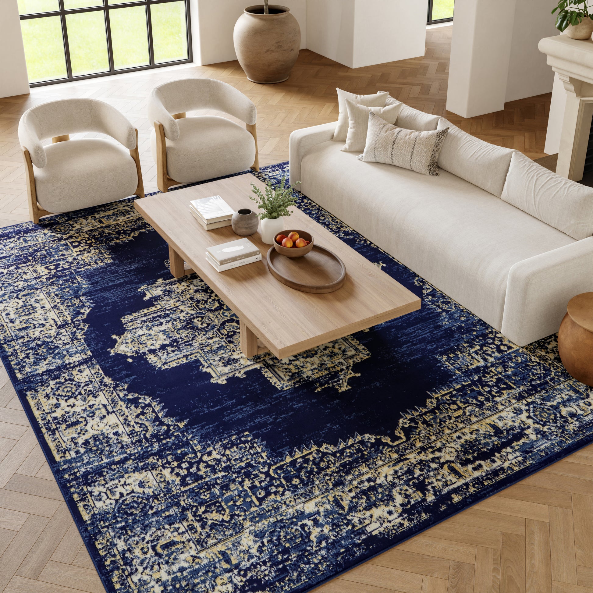 Cream sofa with curved armchairs on a navy distressed rug; light wood coffee table, plants and fruit in a modern living room.