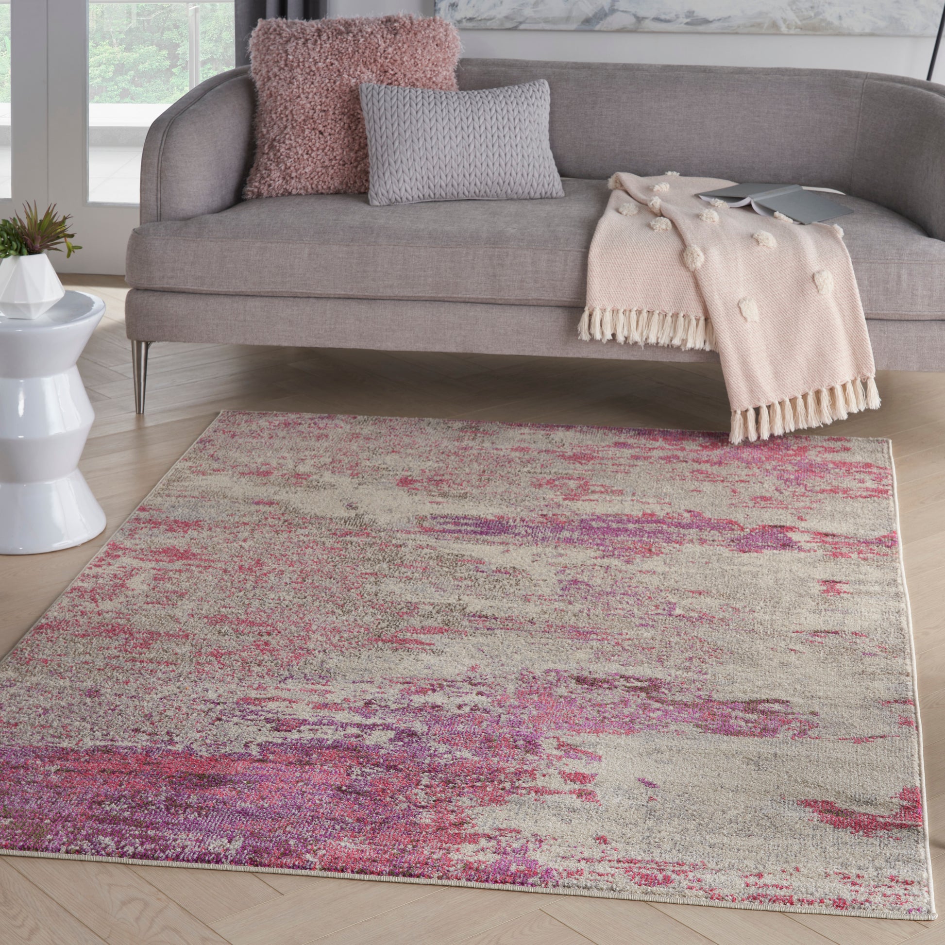 Gray sofa with pink and gray cushions, blush throw blanket, and a beige-pink abstract area rug on light wood floor.