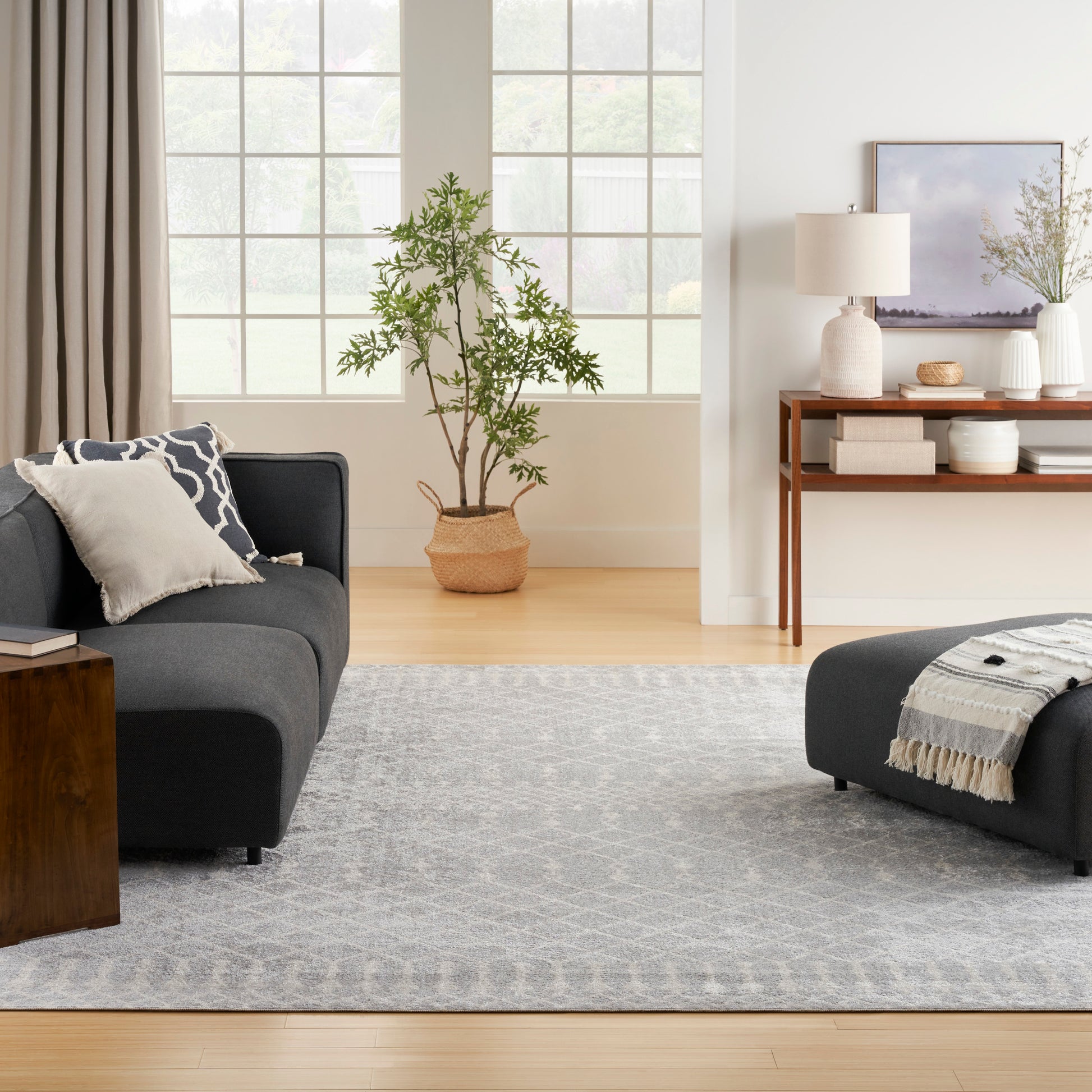 Modern living room with dark gray sectionals, light gray patterned rug, large grid windows, and a potted tree in a wicker basket beside a wooden console.