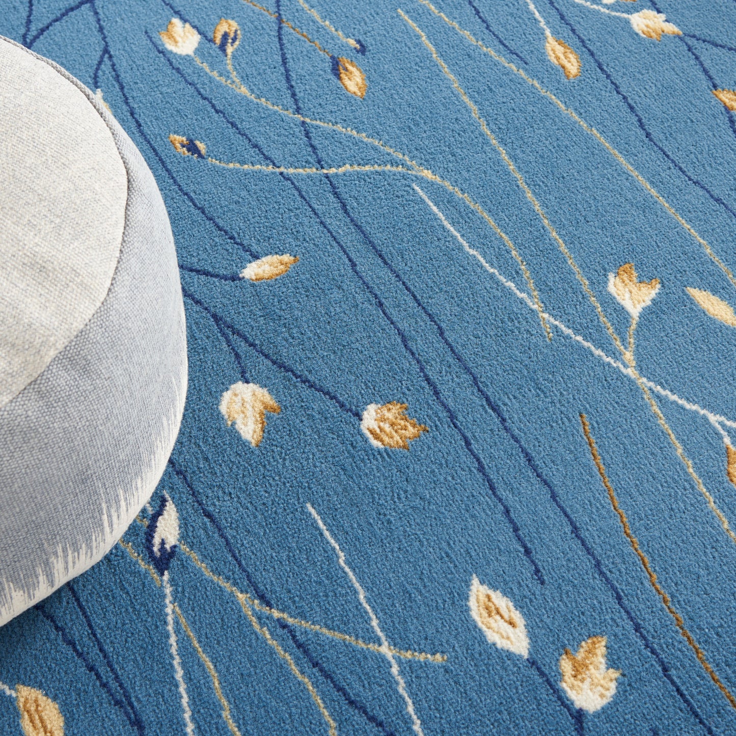 Blue rug with slender white & dark blue lines & gold leaf motifs; round light gray cushion tucked on the edge.