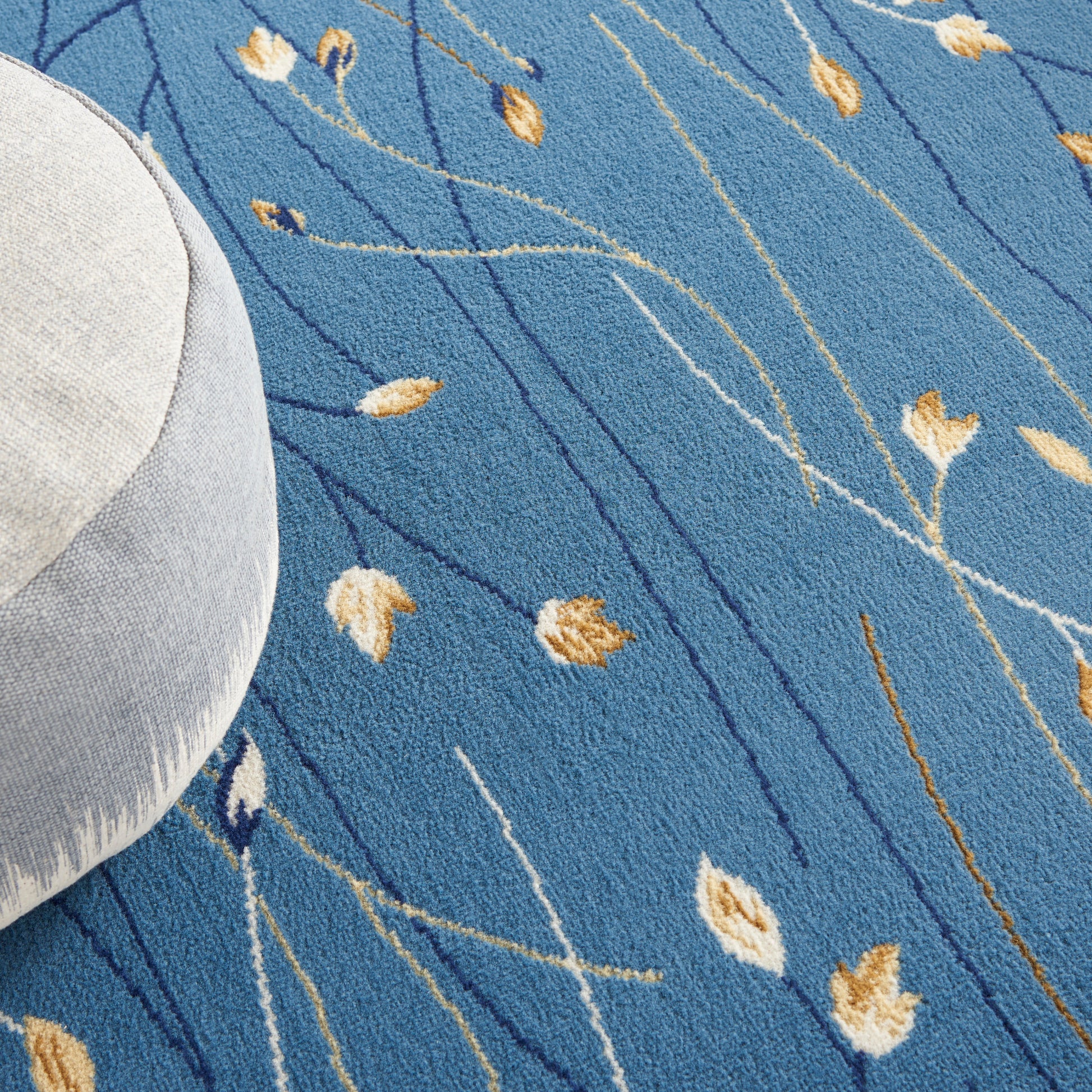 Blue rug with slender white & dark blue lines & gold leaf motifs; round light gray cushion tucked on the edge.