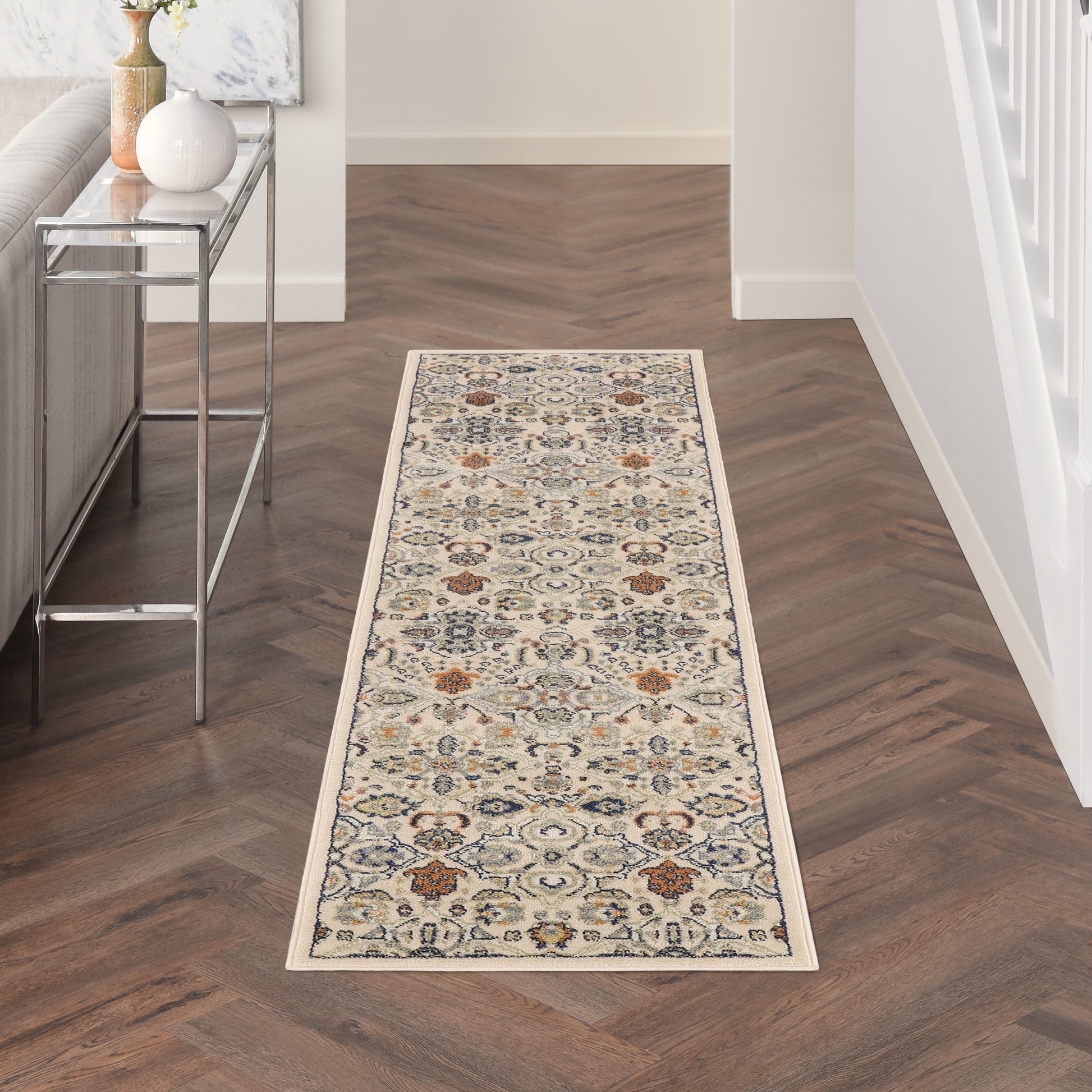 Beige hallway runner rug with blue and rust floral motifs and cream border on dark herringbone wood floor.