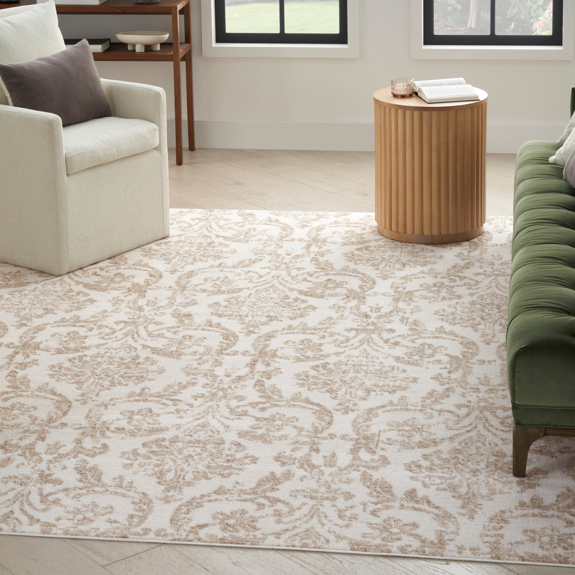 Beige ornate-area rug in a light living room with a cream armchair, green tufted sofa, and round wooden side table.