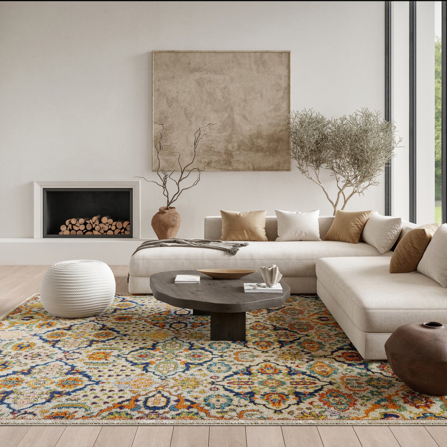 Neutral living room with a beige sectional, round dark-wood coffee table, vibrant patterned rug, white pouf, and potted plants.