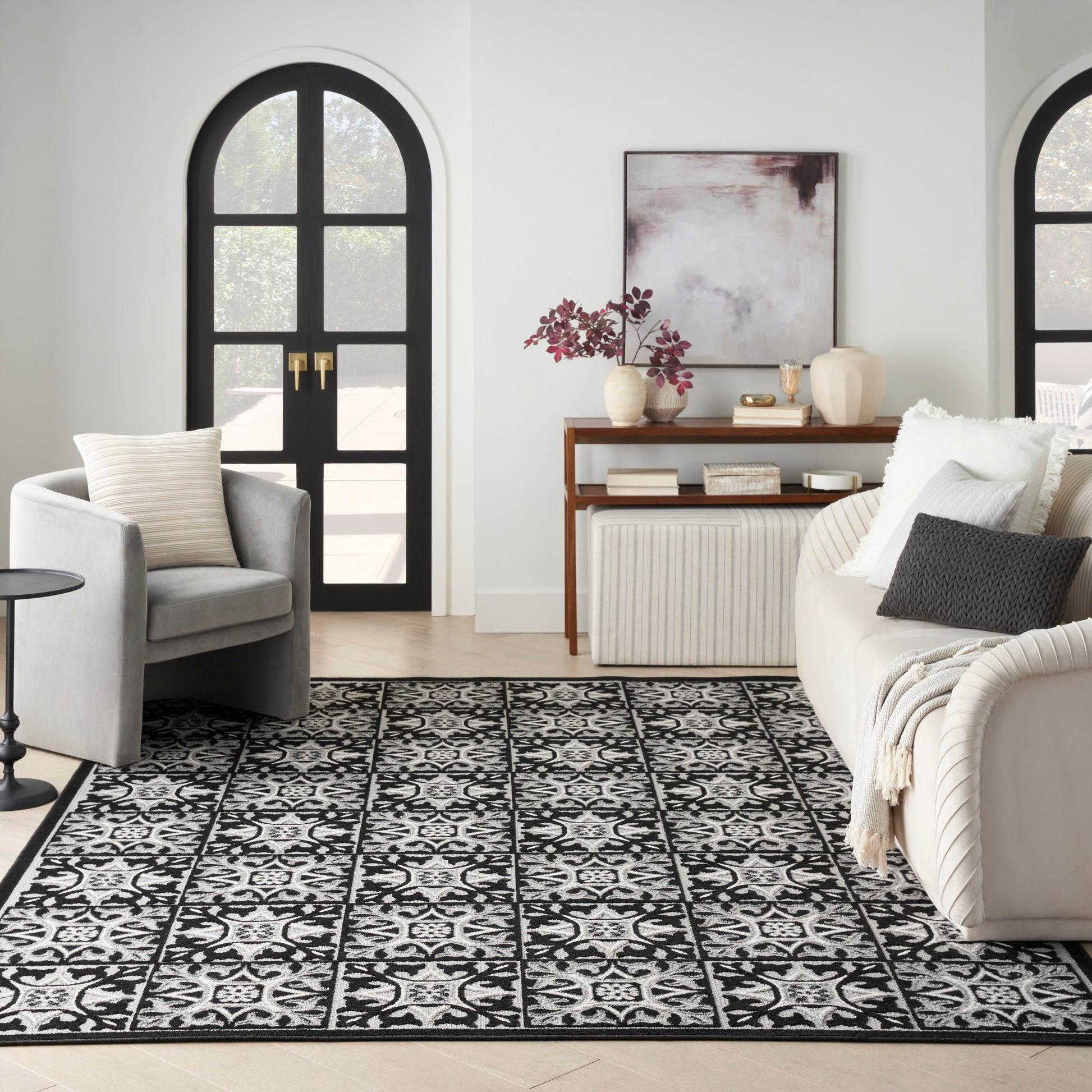 Black & white patterned rug with floral tiles in a living room with gray armchair, beige sofa, and arched black doors.