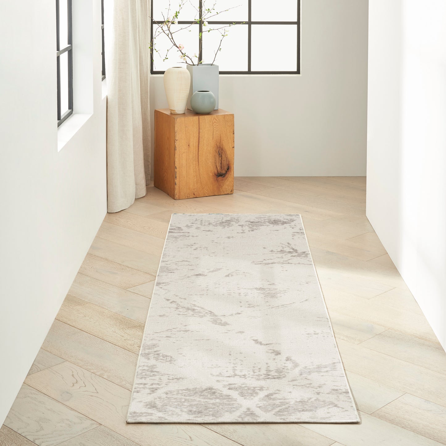 Neutral hallway runner rug with soft gray pattern on light wood floor, wooden side table and ceramic vases by a window.