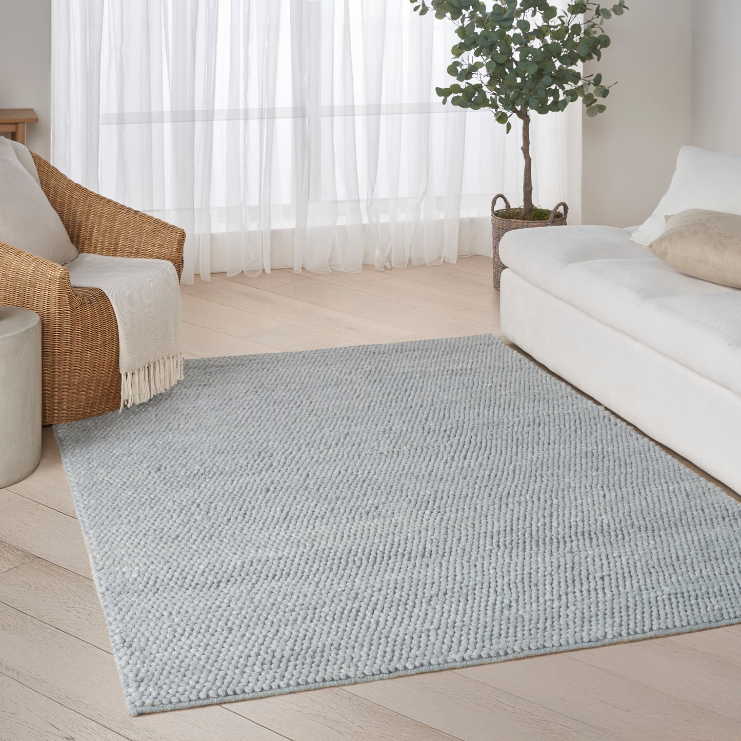 Light gray rectangular textured rug on pale wood floor in a living room with a white sofa, wicker chair, and sheer curtains.
