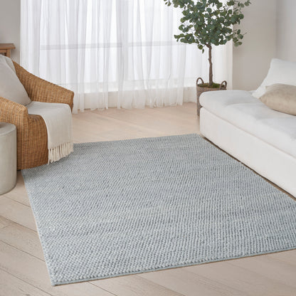Light gray rectangular textured rug on pale wood floor in a living room with a white sofa, wicker chair, and sheer curtains.