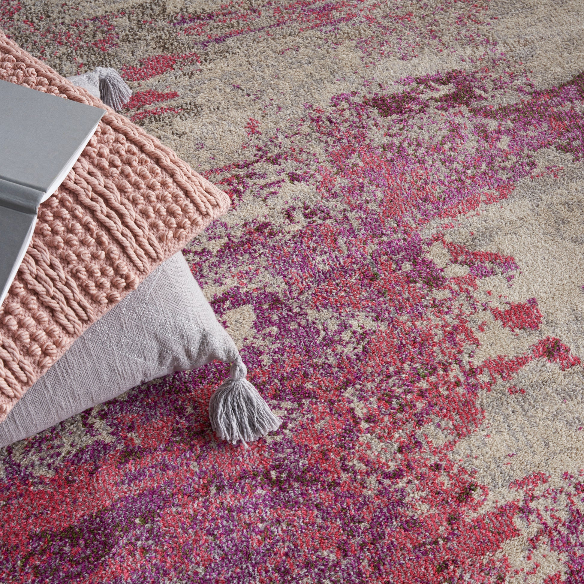 Beige rug with pink and magenta abstract pattern, plus a chunky pink knit pillow and a light gray throw with tassels.