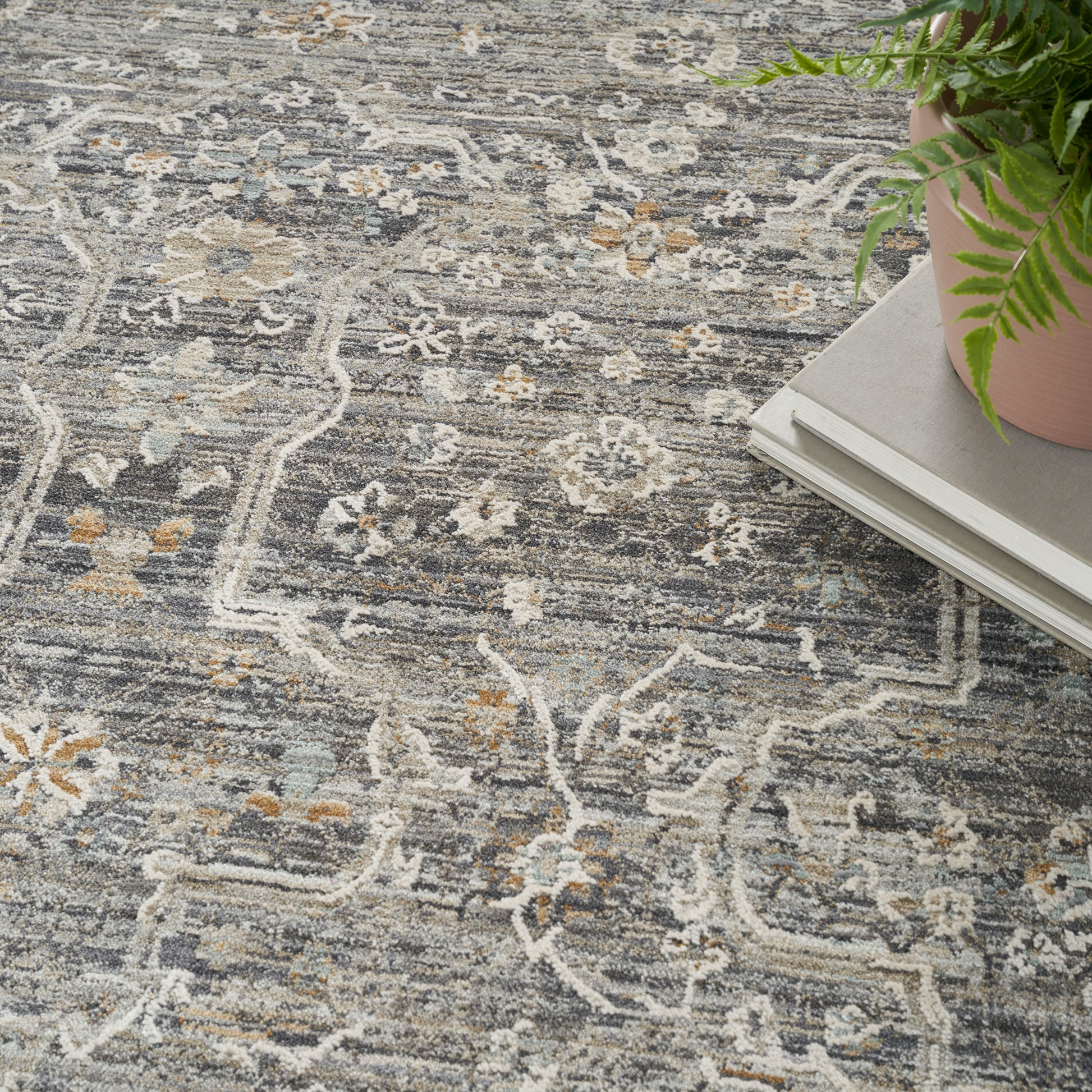 Grey oriental-style rug with white floral motifs and beige accents, corner stack of magazines with a potted fern.