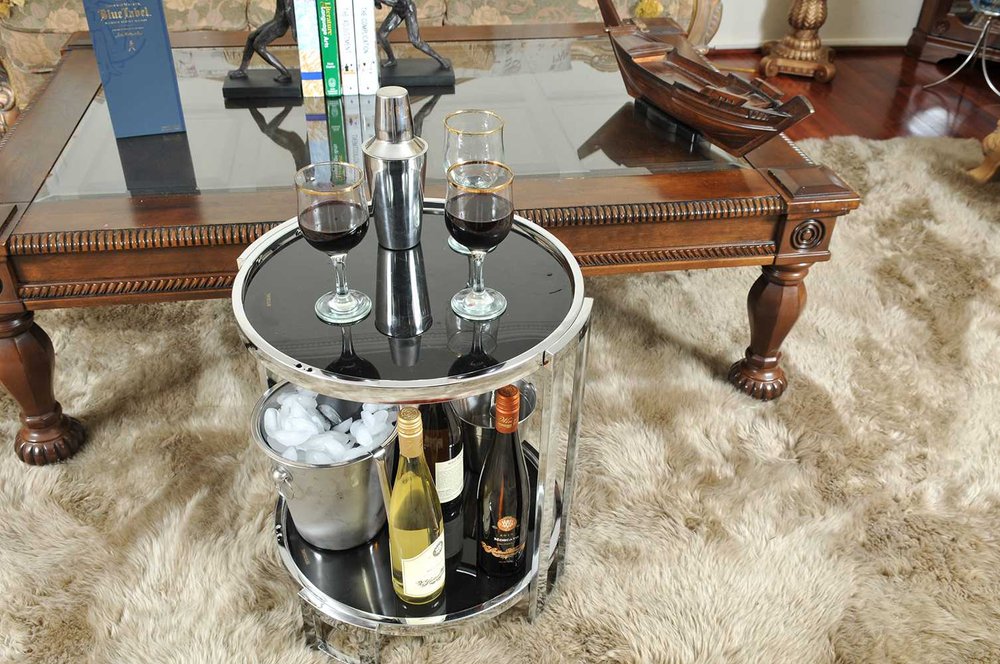 Round chrome two-tier bar cart with glass top; top holds two wine glasses and shaker, bottom stores wine bottles and ice tub.