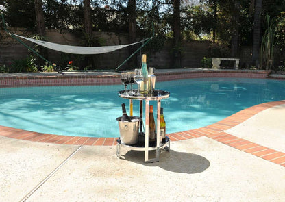 Three-tier stainless steel bar cart with wine bottle, glasses and ice bucket beside a blue pool on brick patio.