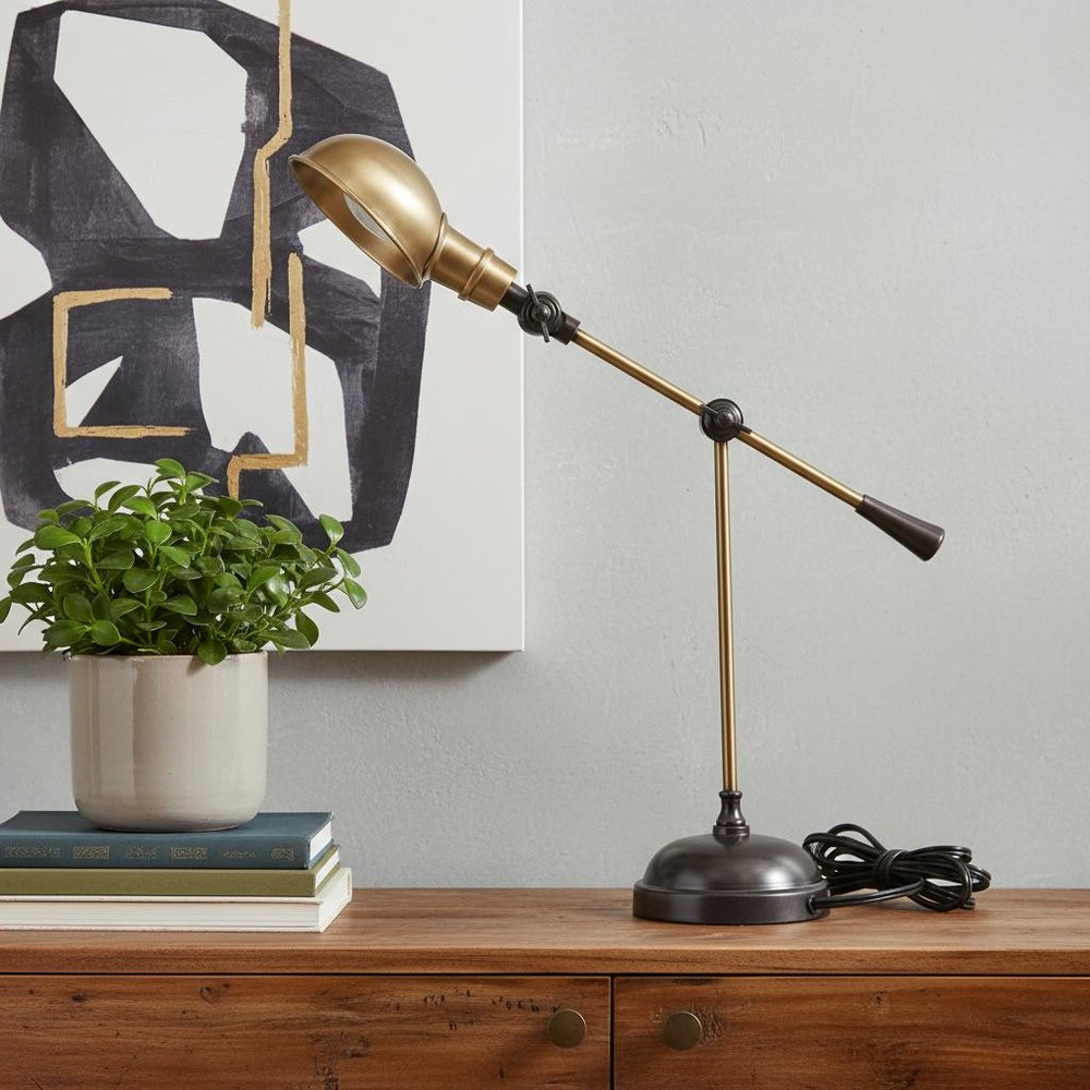 Brass adjustable desk lamp with black joints on a wooden desk beside a potted plant and stacked books.