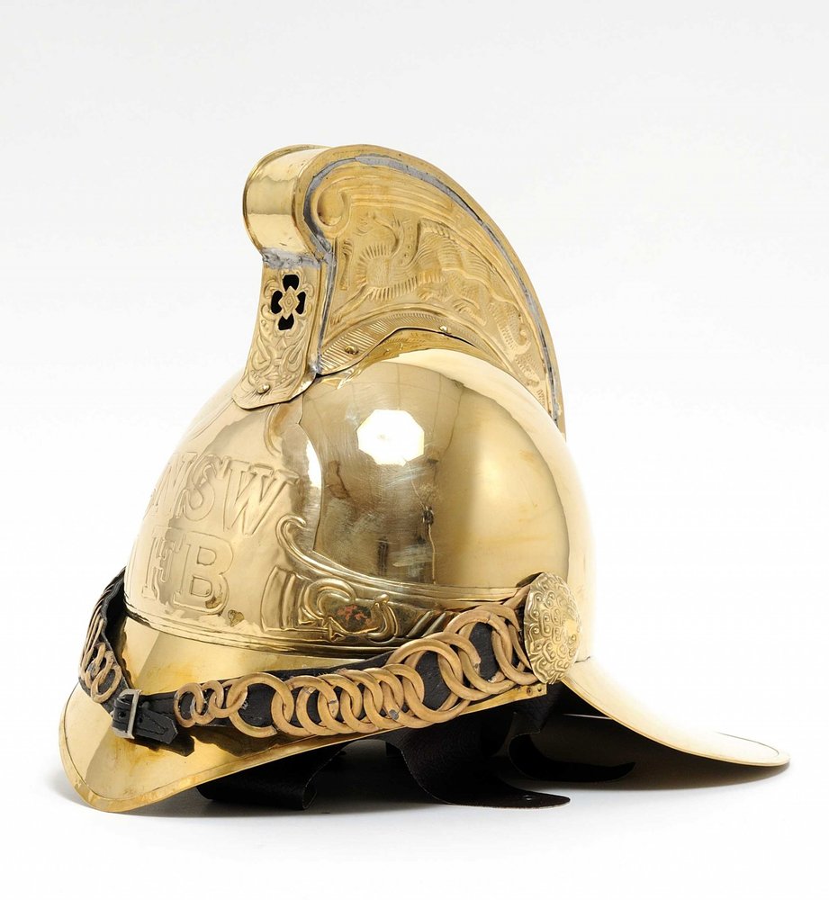 Polished brass firefighting helmet with engraved crest, ornate detailing, chain trim, and leather chin strap.