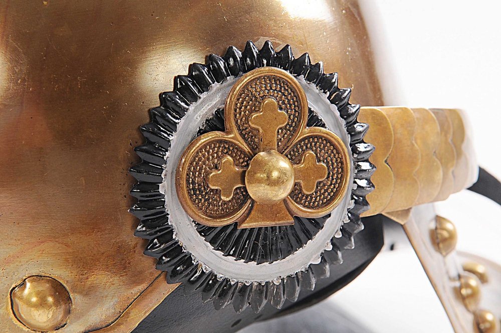 Brass helmet detail with black cog gear, gold quatrefoil centerpiece and leather strap rivets; steampunk style.