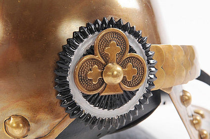 Brass helmet detail with black cog gear, gold quatrefoil centerpiece and leather strap rivets; steampunk style.
