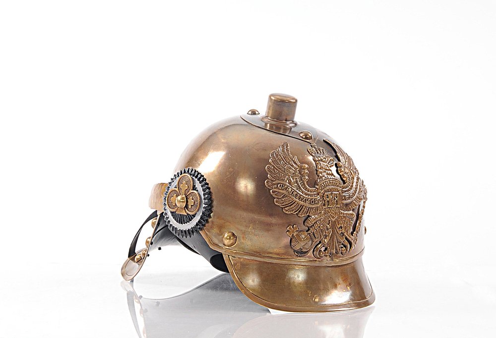Brass-toned ceremonial helmet with embossed imperial eagle, black leather strap, and decorative rosette on the side.