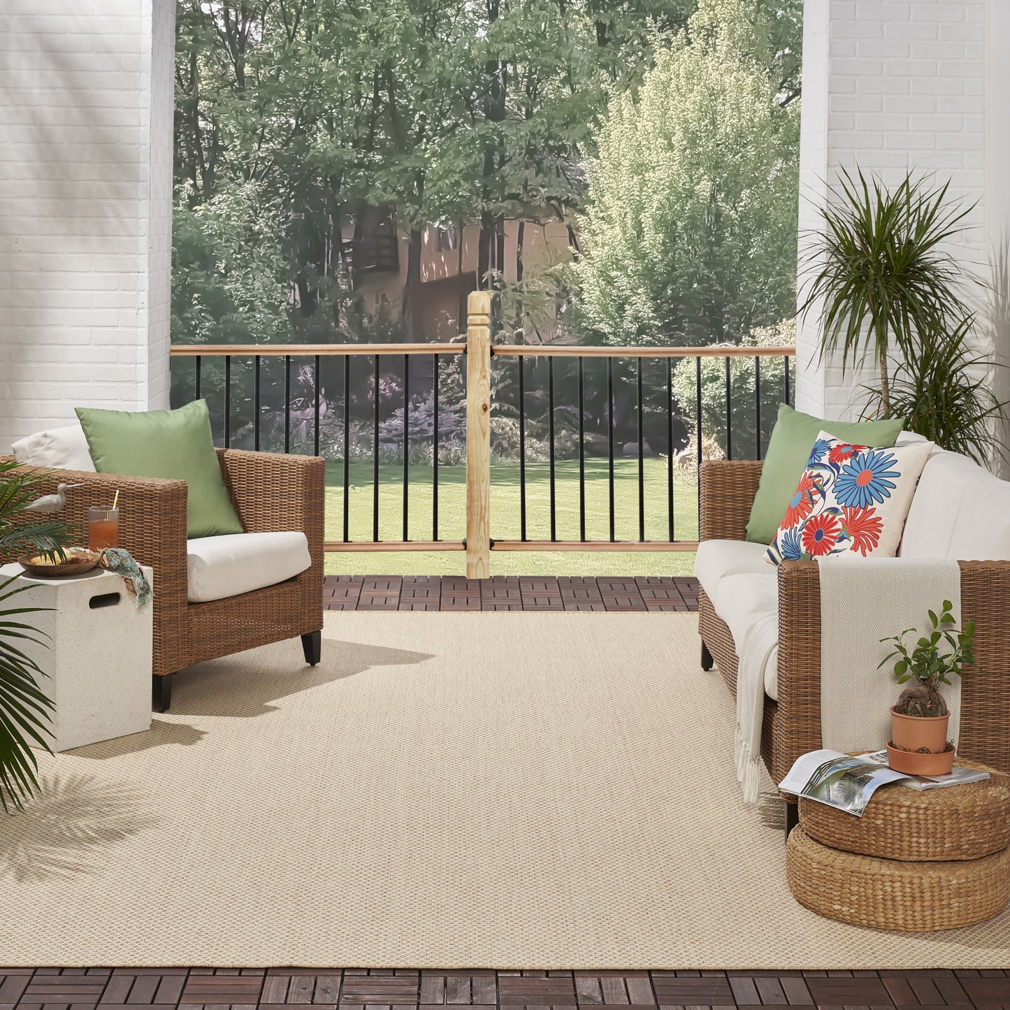 Wicker outdoor seating on a patio with beige rug, white cushions, green and floral pillows, potted plants, and wooden railing.