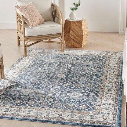 Blue and ivory patterned rug on light wood floor; rattan chair with beige cushion near a geometric wooden side table.