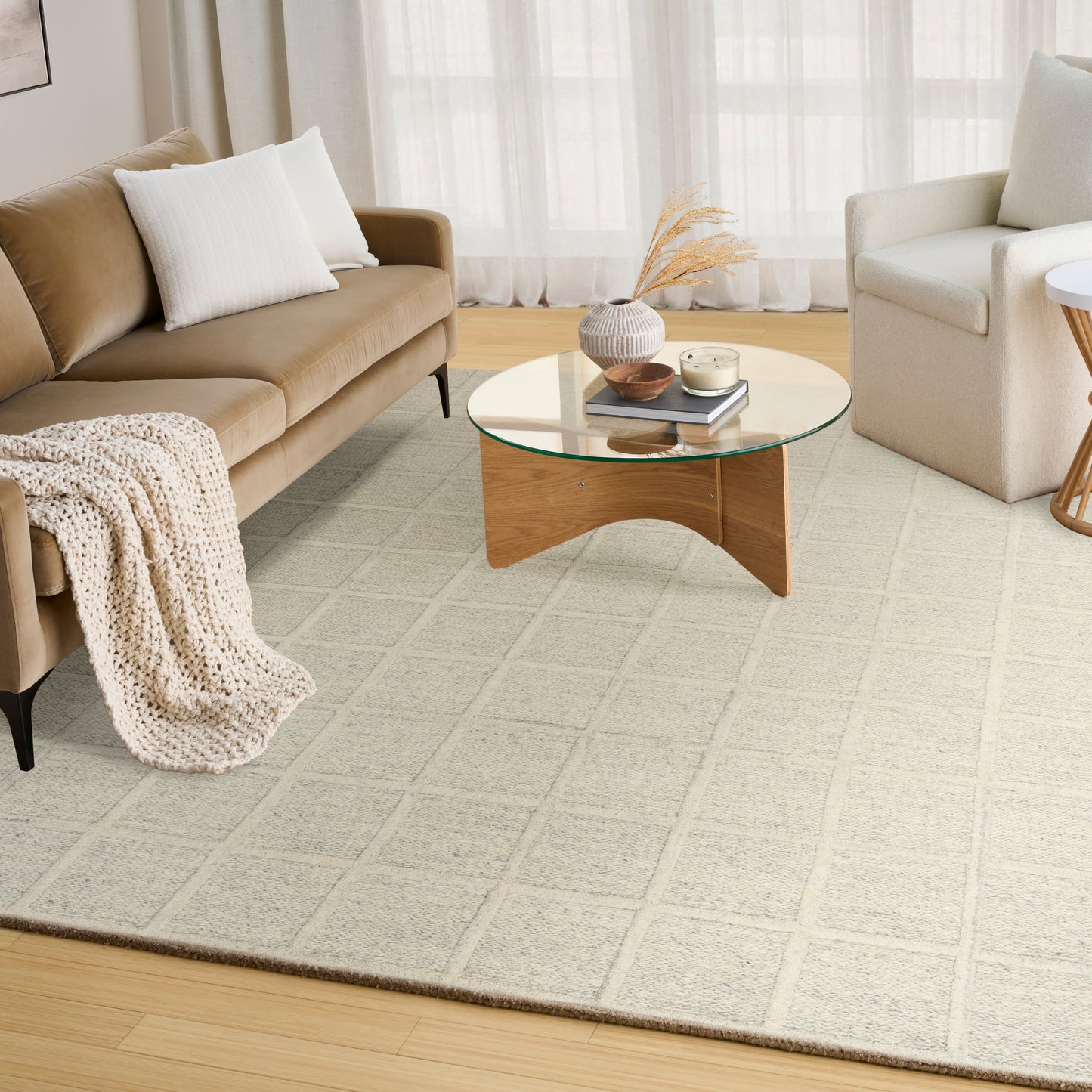 Living room with tan sofa, white pillows, cream armchair, glass-top round coffee table and beige grid rug