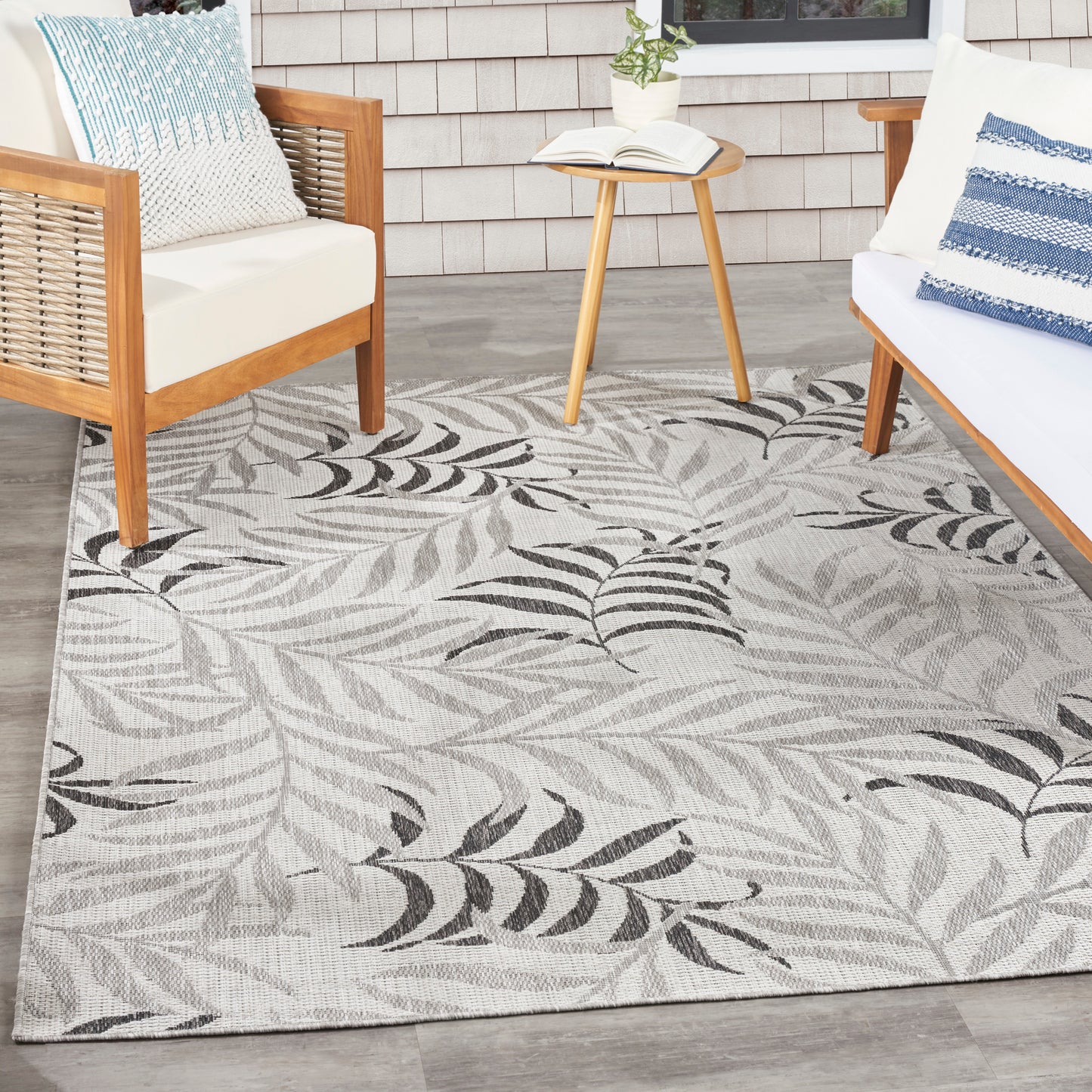 Outdoor rug with gray background and black fern leaves; wooden chairs, matching cushions, small side table with plant.