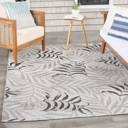 Outdoor rug with gray background and black fern leaves; wooden chairs, matching cushions, small side table with plant.