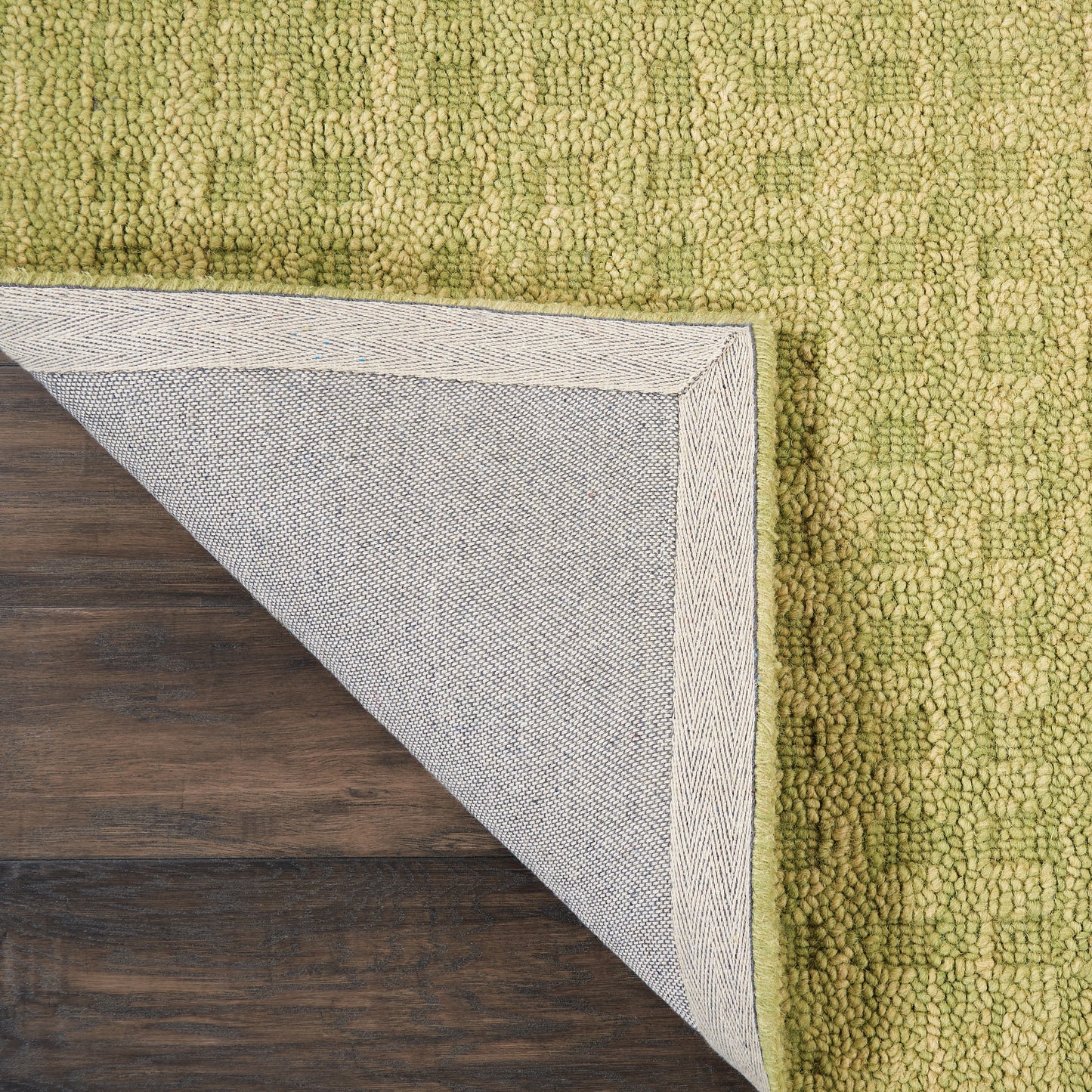 Green chunky-textured rug with loop pile; folded corner reveals gray backing with woven border on wood floor.