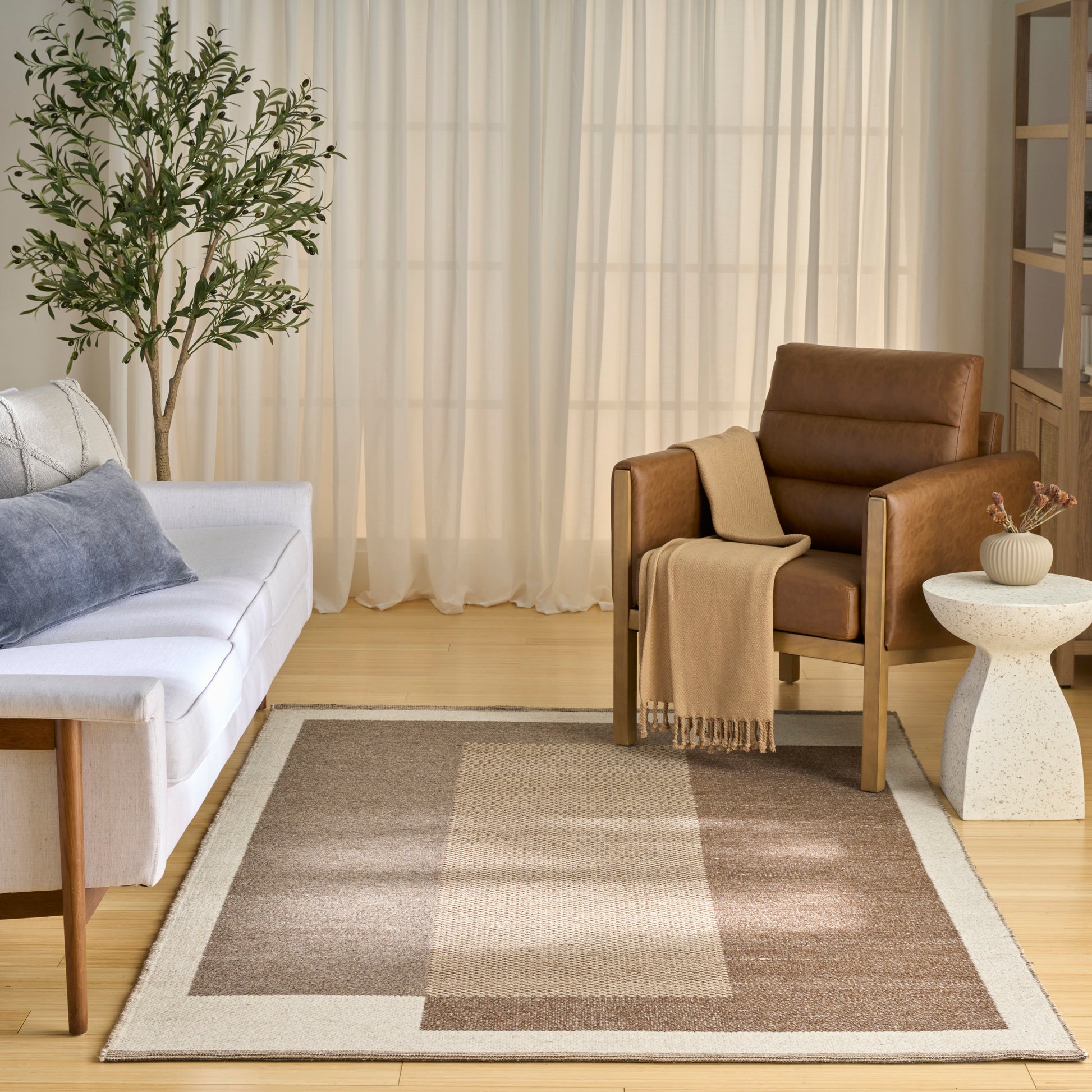 Living room with beige-brown geometric area rug, white sofa, brown leather armchair, throw blanket, plant, and round table.