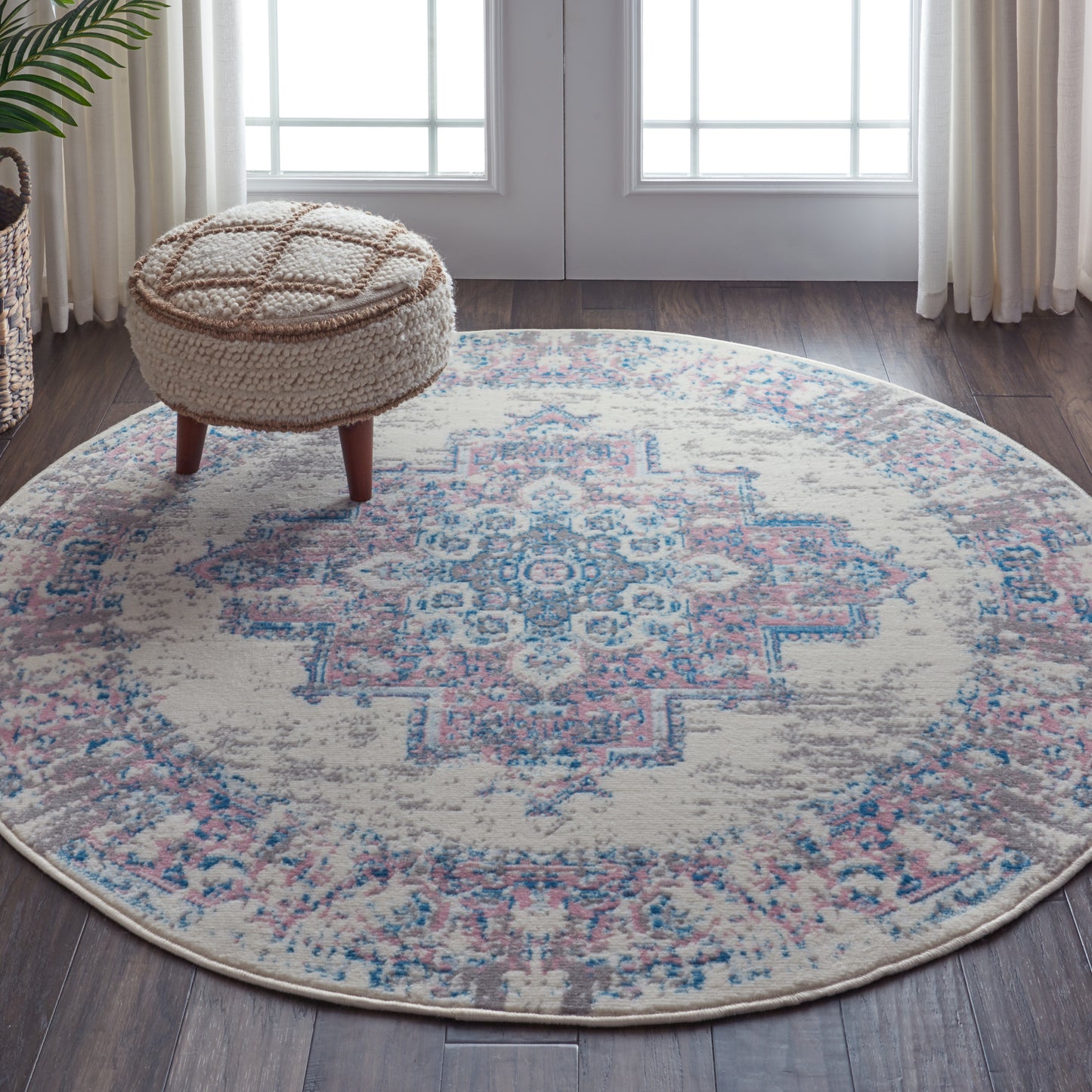 Round pastel rug with cream, blue and pink geometric medallion, beside a beige knitted pouf with wooden legs.