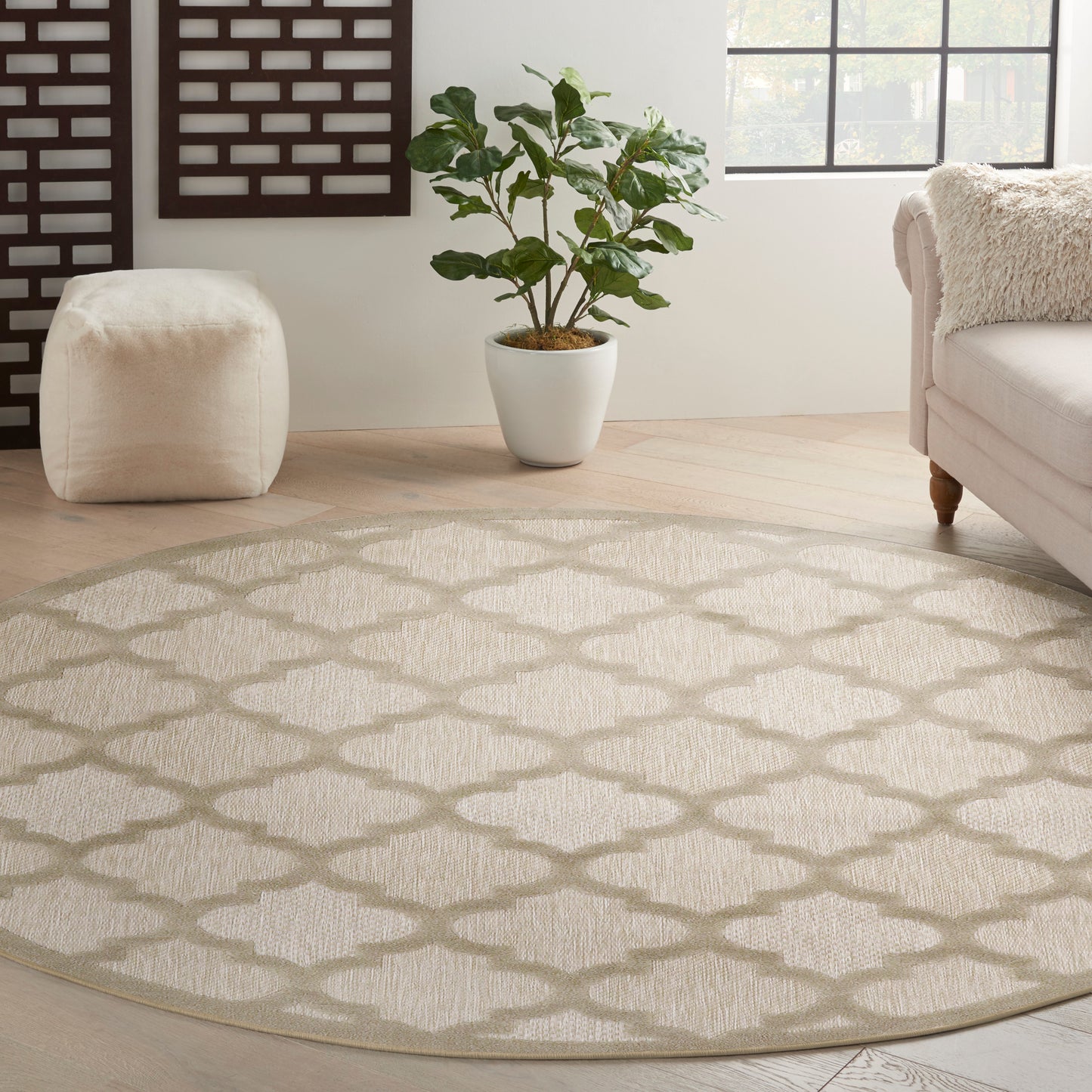 Round beige woven area rug with a diamond lattice pattern in a neutral living room with a plant, pouf, and sofa.