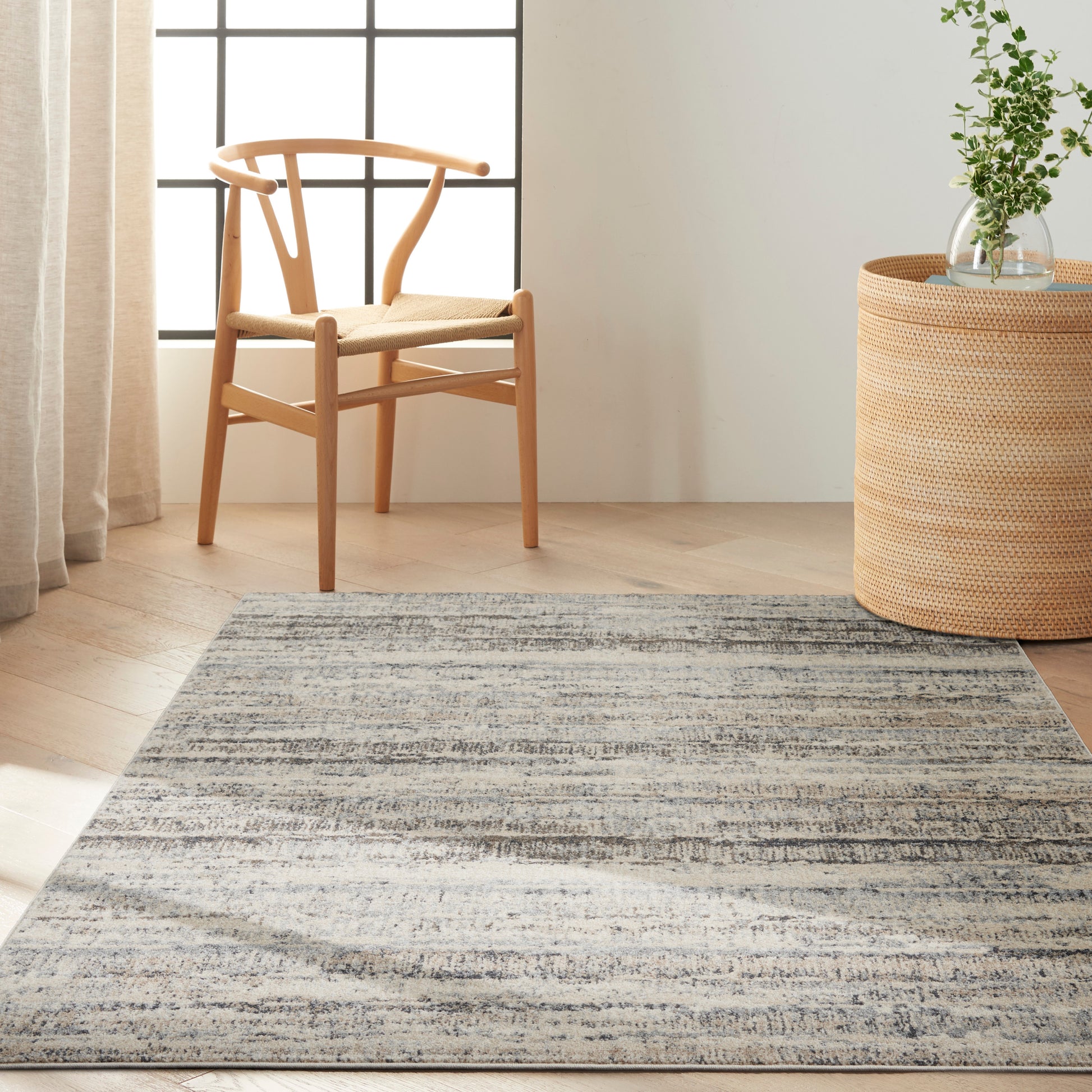 Light oak chair with woven seat beside a round wicker side table holding a glass vase of greenery on a blue-gray striped rug.