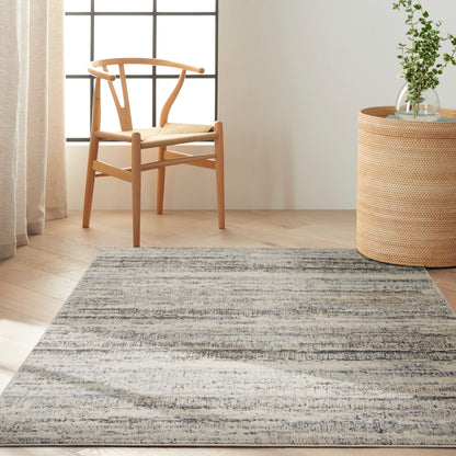 Light oak chair with woven seat beside a round wicker side table holding a glass vase of greenery on a blue-gray striped rug.