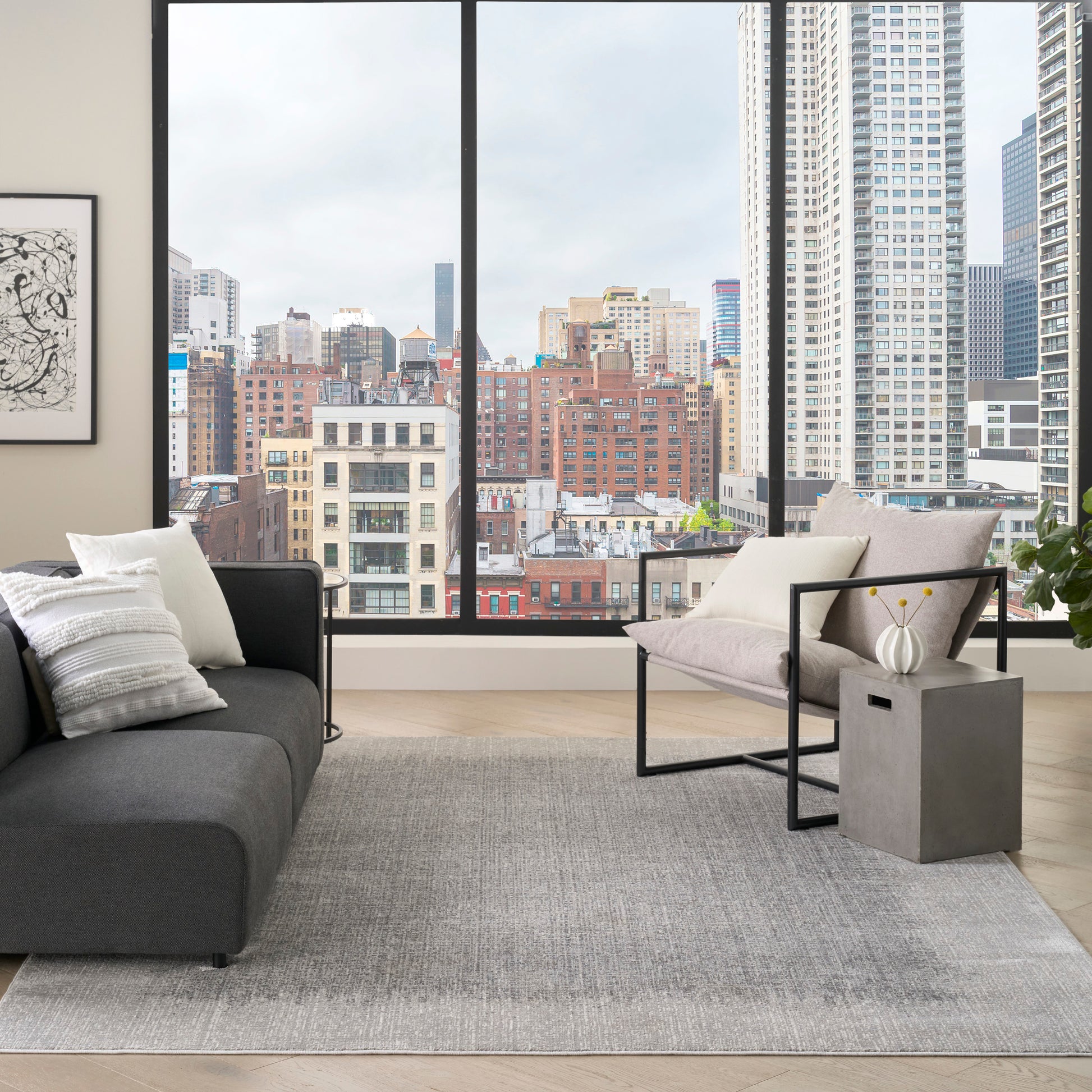 Modern living room with large windows overlooking city skyline; dark gray sofa, light rug, metal-framed chair, concrete side table, plant.