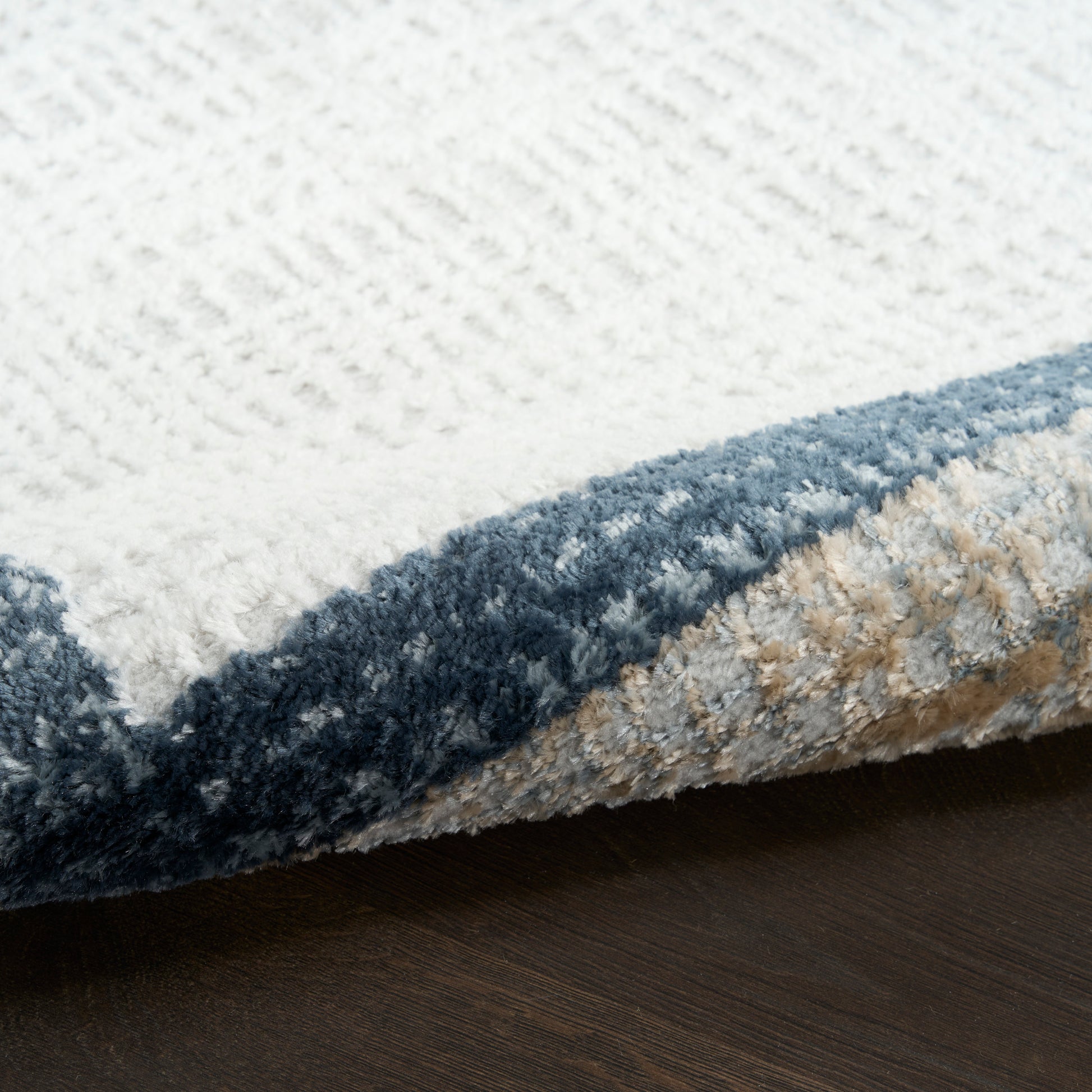 Close-up of a thick, textured rug with a white field and blue border, folded to reveal beige underside on dark wood floor