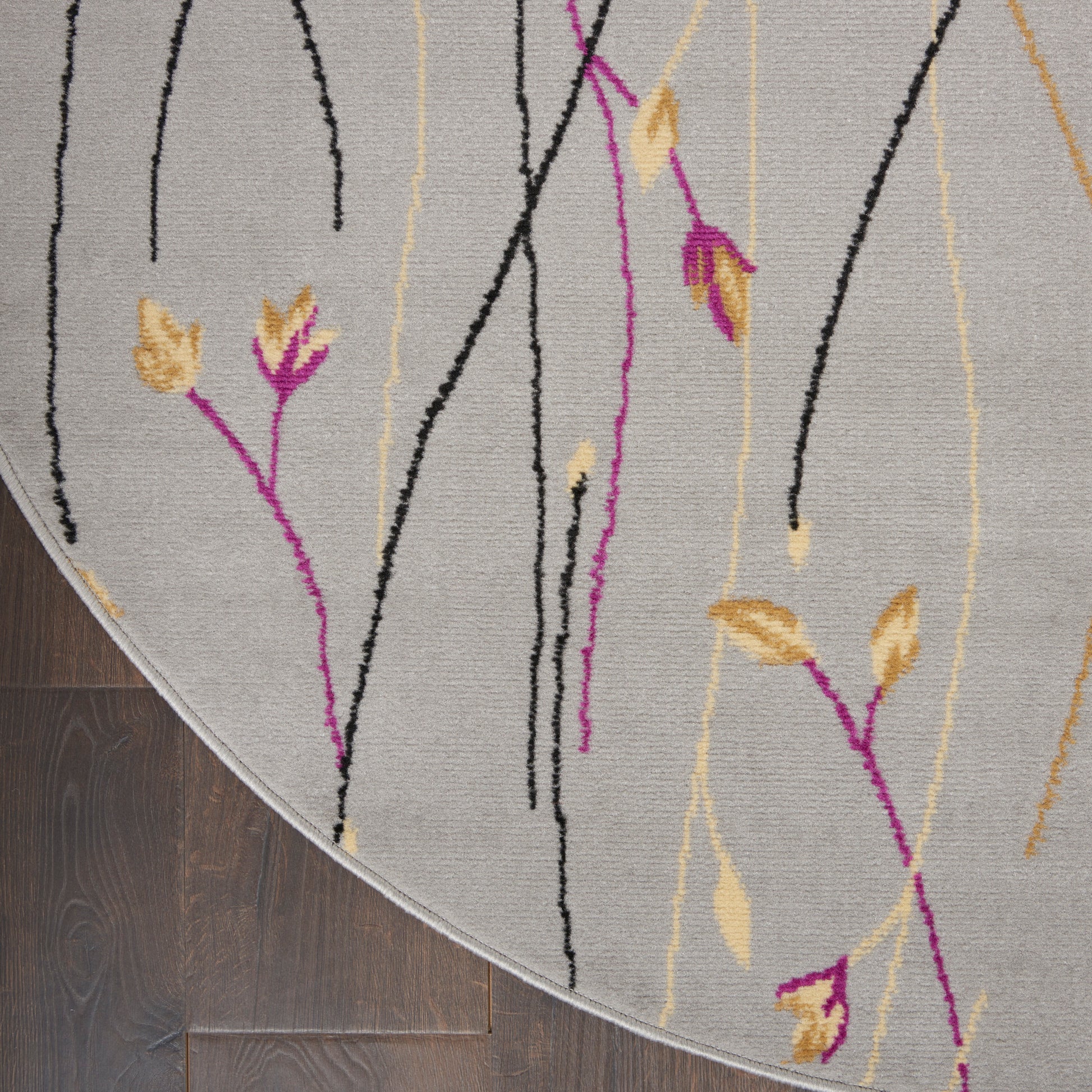 Round grey rug with abstract black and yellow vertical stems and magenta leaf accents on dark wood floor.